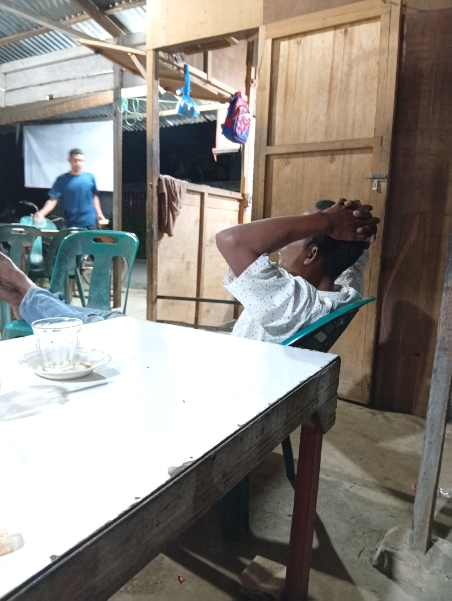 This image depicts a casual scene, likely in Lhokseumawe, Indonesia, during the evening or night, indicated by the artificial lighting and dark surroundings.
The primary subject is a man sitting in a green plastic chair, leaning back with his hands clasped behind his head. He is wearing a white patterned short-sleeved shirt and appears relaxed, possibly contemplating or resting. In the foreground, a white table holds a clear glass with a small amount of liquid and a saucer. The table has a rough wooden edge. To the left of the main subject, another person, a man wearing a blue t-shirt, is visible in the background. He is standing near a motorcycle, suggesting a casual gathering or waiting area.
The setting appears to be an open-air or semi-open structure with a corrugated metal roof supported by wooden beams. Wooden walls and doors are visible in the background, contributing to a rustic or utilitarian atmosphere. The lighting is dim, casting shadows and highlighting the textures of the wood and corrugated metal. A blue object, possibly a bag or toy, hangs from a beam.
There is no visible text in the image. The overall mood is one of quiet repose and informal social interaction.
