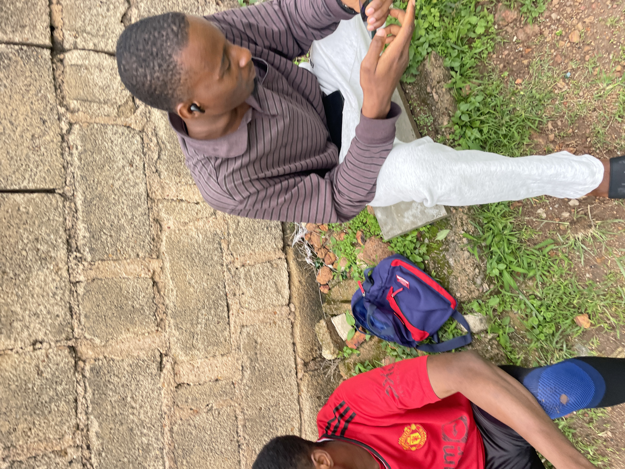 Two young men are seated outdoors next to a brick wall. The man on the left wears a red Manchester United jersey with black and white stripes on the sleeves and a blue knee brace. The man on the right wears a brown and grey striped long-sleeved shirt and light grey pants. He has black wireless earbuds in his ears and is looking down at his hands, which are holding a smartphone. A blue and red backpack rests on the ground between the two men. The ground is covered in dirt, grass, and small rocks. The scene is consistent with a casual outdoor setting in the city of Jos, Nigeria.