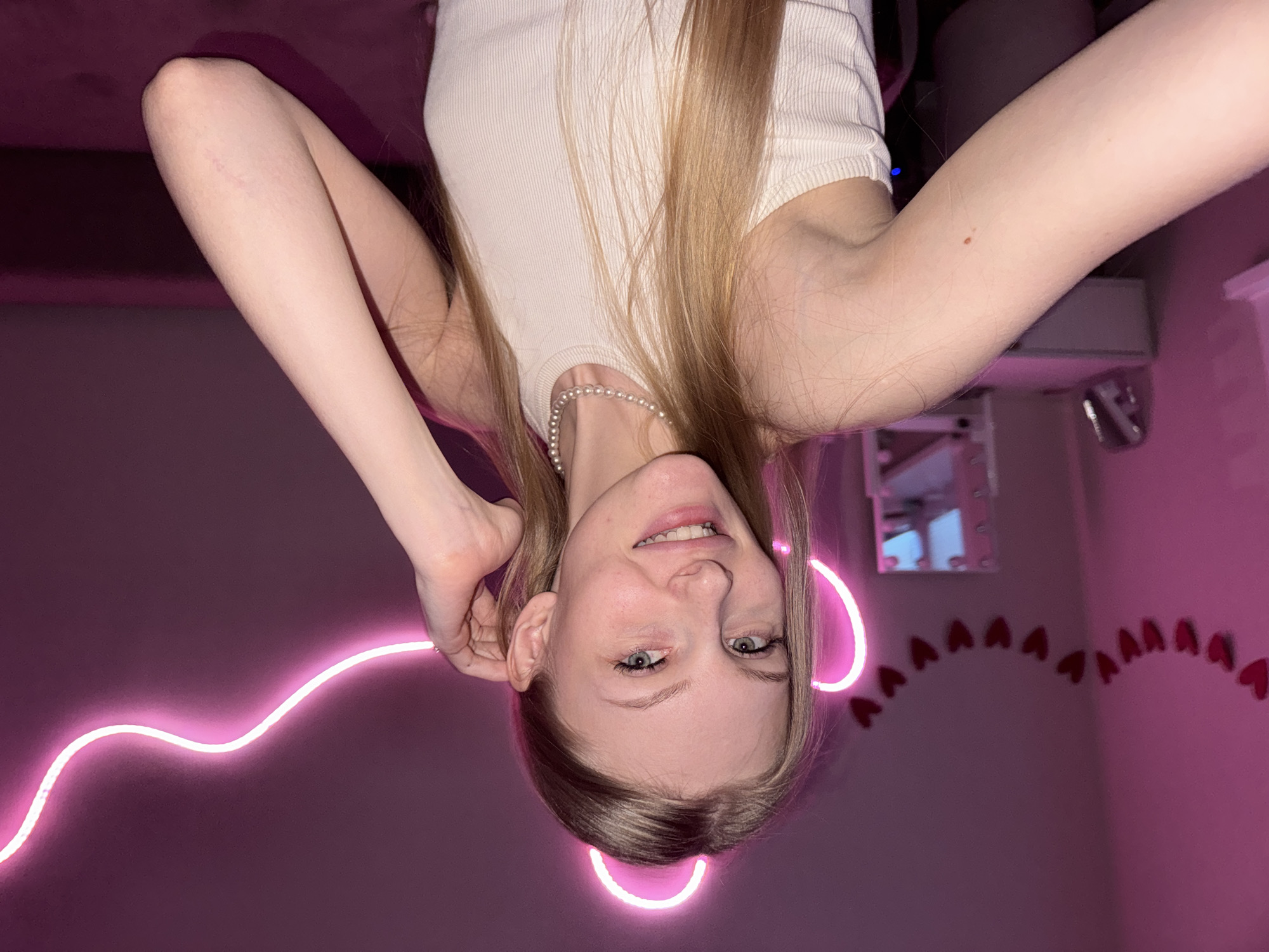 A young fair-skinned female with long, light-colored hair and blue eyes is depicted, smiling and looking directly at the camera. She is wearing a light-colored, possibly white, ribbed tank top and a pearl necklace. Her right arm is bent with her hand positioned near her head. A faint linear mark is visible on her right elbow, and a small red circular mark, along with some faint scratches, is present on her right forearm.
The scene is an indoor space, predominantly illuminated by prominent pink neon lights. A wavy strip of neon light extends diagonally across the wall behind her. Additionally, two curved neon strips form ear-like shapes above the subject's head. Below the subject's head, a vertical line of small red heart-shaped decorations is affixed to the wall. In the background, a reflective surface, possibly a mirror, shows a blurred reflection of other room elements, including what appears to be a white computer tower and a dark chair. The dominant color scheme of the environment is shades of pink and magenta due to the ambient lighting.
There are no discernible visual cues within the image to indicate the specified location of Perm, Russia.