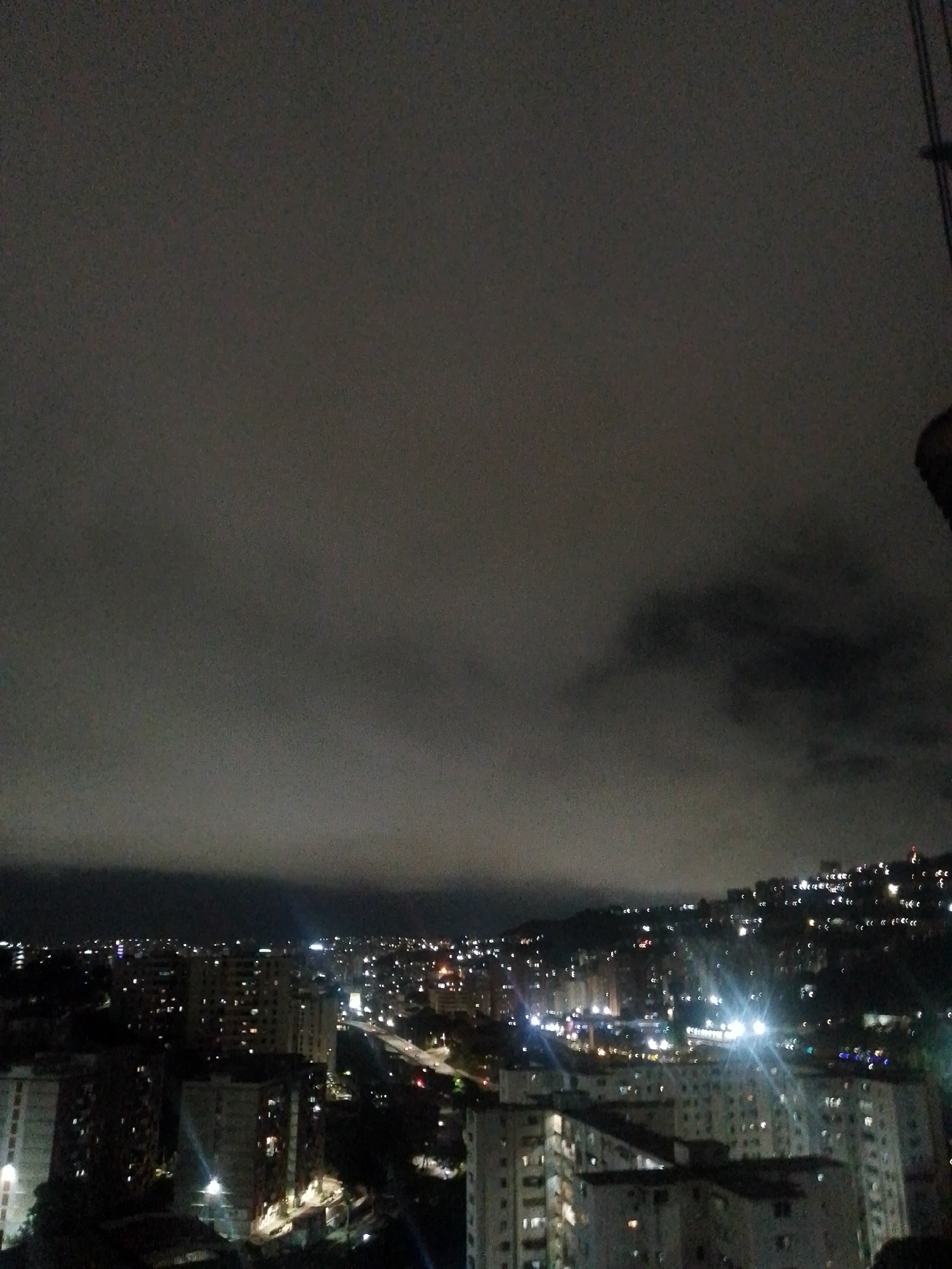 The image shows an aerial view of a densely populated urban area at night, likely in Caracas, Venezuela, given the hilly terrain and architectural style. The foreground features several large apartment buildings, with numerous illuminated windows indicating occupied residences. In the middle ground, more buildings are visible, interspersed with roads and streetlights that create streaks of light due to the long exposure. The background reveals a hillside covered with smaller dwellings and lights, suggesting a spontaneous or informal settlement climbing the slopes. The sky is overcast with dark clouds, obscuring any stars or moon, and the overall lighting is dim, with the city's artificial lights being the primary source of illumination. There are no people or animals clearly visible in the frame. The scene depicts a sprawling cityscape at night, characterized by numerous residential structures and the ambient glow of urban life under a cloudy sky.