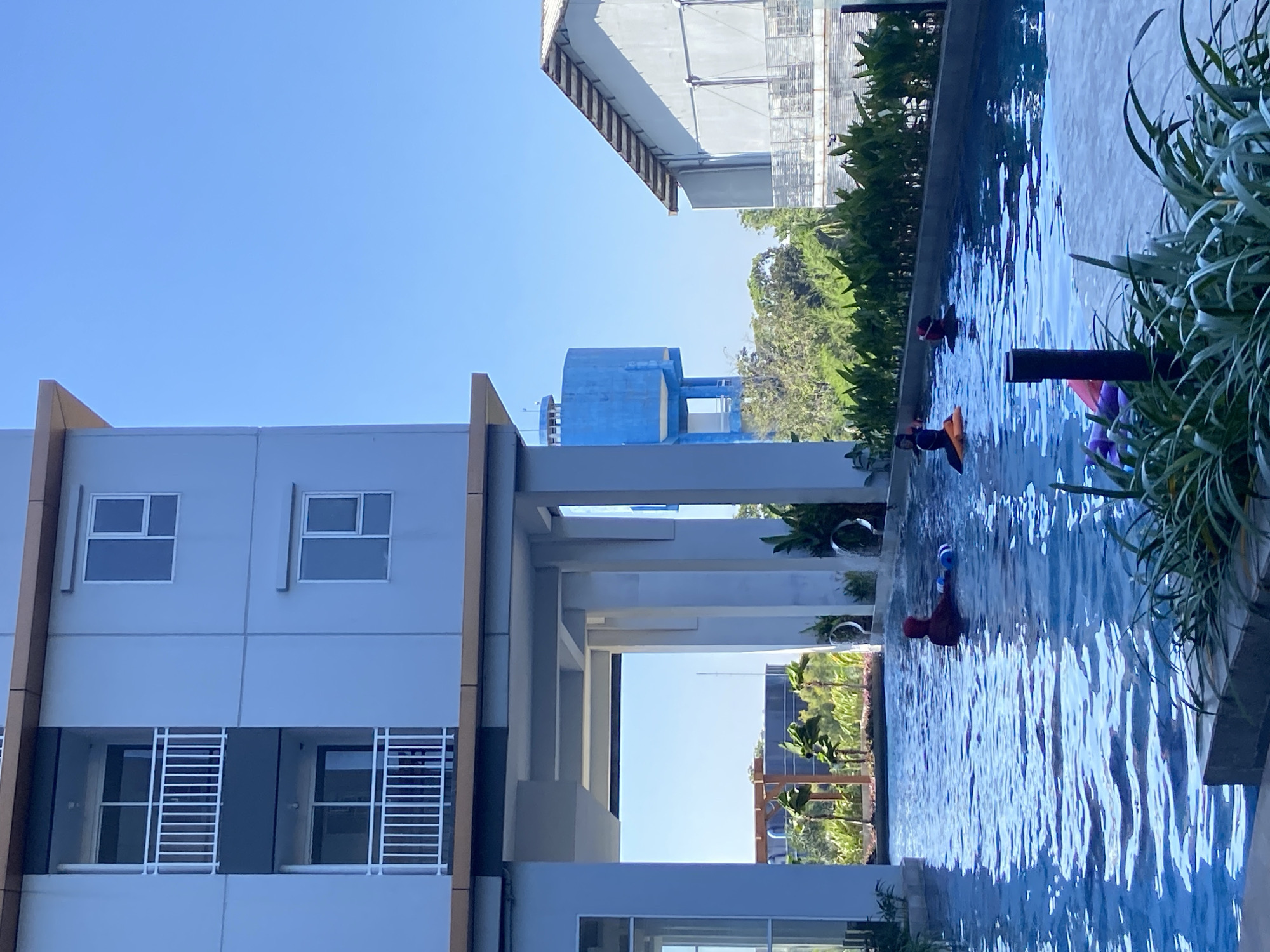 In Sidoarjo, Indonesia, a swimming pool is situated beside a multi-story building. Several individuals are in the pool, some with inflatable devices. The water is calm and reflects the clear blue sky. To the left of the pool is a light blue building with balconies. In the background, a blue water tower is visible, along with lush green trees and another building. The foreground is partially obscured by green plants.