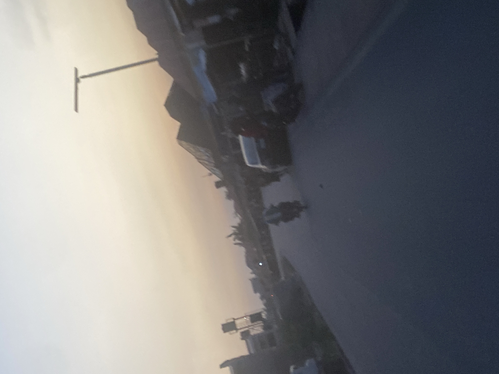 The image depicts a street scene in Abuja, Nigeria, likely at dusk or dawn, based on the dim, muted light. The view is slightly tilted and captures a portion of a street with buildings and vehicles lining it.
**Environment:**
* The sky is a gradient of pale yellow and light blue, suggesting either sunrise or sunset.
* The street surface appears wet, possibly from rain or street cleaning.
**Objects:**
* A utility pole stands prominently against the sky.
* Buildings of varying heights are visible along the street, mostly with indistinct features due to the light.
* Cars and motorcycles can be identified parked or moving along the road.
**People:**
* There is no information on people in this picture.
