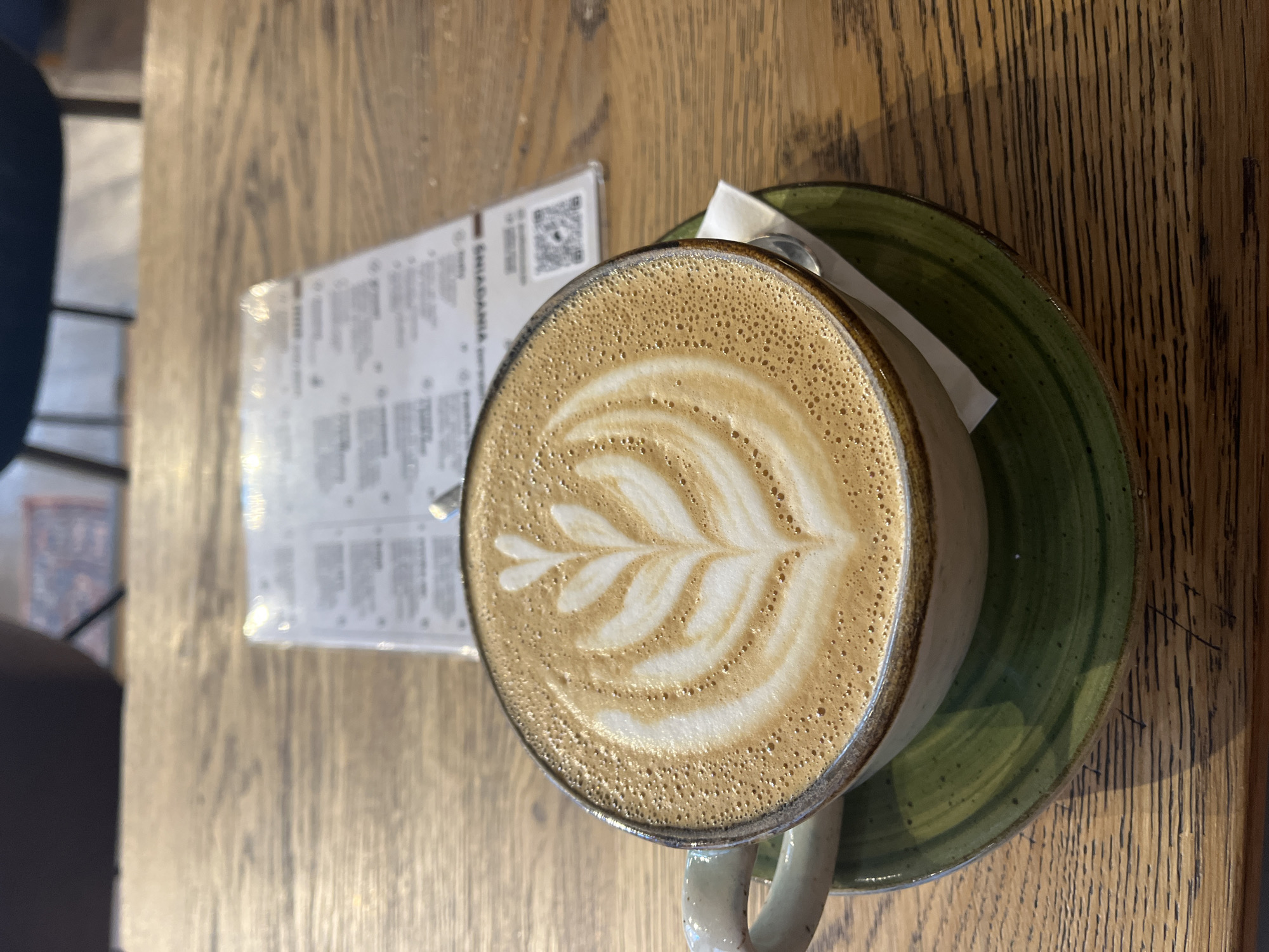 The image depicts a coffee drink with latte art, served on a green saucer, placed on a wooden table. Beside it is a partially visible menu. The location is Unknown, Unknown.
