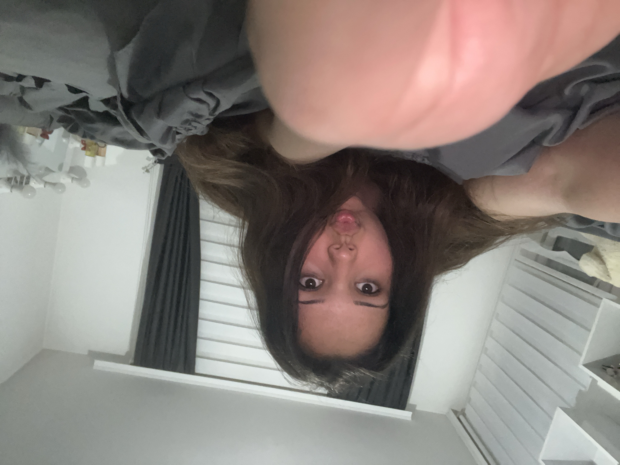 The image is a selfie taken by a young female lying on a bed with grey sheets. She has long, dark brown hair and is making a kissing face with her lips pursed. Her eyes are open and looking towards the camera. In the foreground, her right thumb is blurred and prominently positioned. The background shows a white wall with horizontal blinds, dark grey curtains, and a shelf unit on the left. The lighting suggests an indoor setting, possibly a bedroom. There is no discernible information about the location being Zlatograd, Bulgaria, or any specific city context.