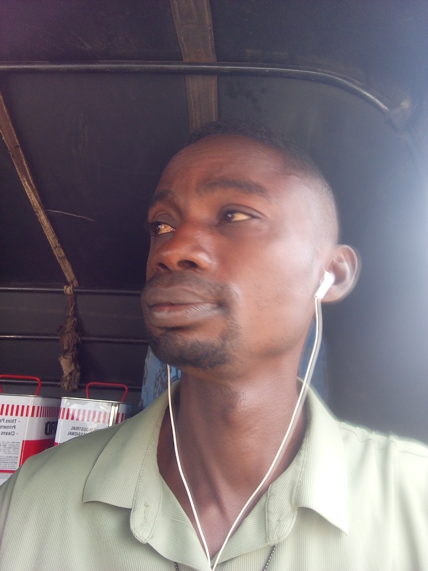 A close-up, shoulder-up, medium shot captures a man with dark skin and a close-cropped haircut, wearing a light green collared shirt and white earbuds. He is looking slightly to his right, his left eye visible. A thin chain necklace is visible around his neck. Behind him, the dark interior of a vehicle is visible, with wooden beams and straps. In the background, to the left, two red-topped cylindrical containers with white labels are partially visible. The image appears to have been taken inside a vehicle, possibly a motorized tricycle, given the structural elements in the background. The lighting suggests it is daytime, with a bright light source coming from the right.