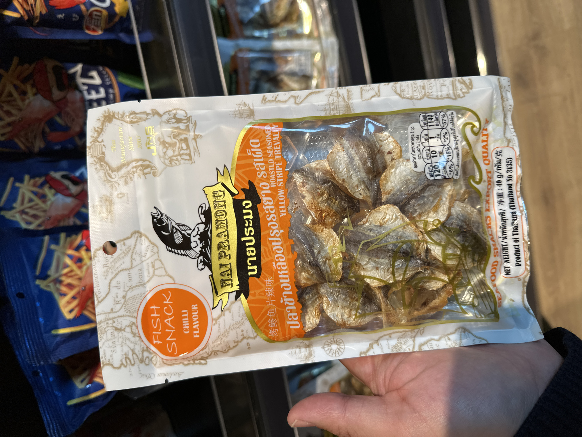 A person's hand holds a white and transparent package of "Nai Pramong" fish snack, chilli flavour. The packaging shows a map-like design, a black fish logo, and nutritional information in Thai script. The snack appears to be dried fish, visible through the clear plastic section. In the background, blurred shelves suggest a grocery store setting, with other snack packages visible.