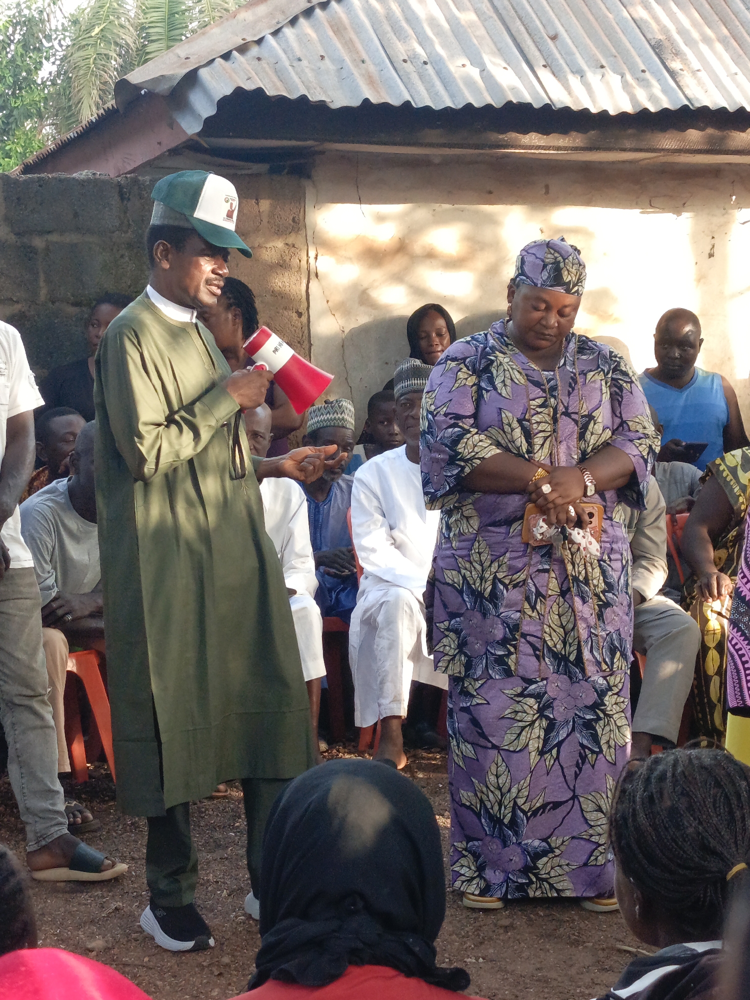 This is a medium-shot, eye-level photograph taken outdoors in what appears to be a rural or semi-urban setting in Akwanga, Nigeria. The scene depicts a gathering of people, possibly a community meeting or event.
In the foreground left, a man wearing a green robe and a green and white baseball cap is holding a red and white megaphone and appears to be speaking. He has a mustache and is looking towards his right. Behind him and to the right, a woman is standing, wearing a purple and blue patterned dress and a matching headwrap. She is looking down with her hands clasped in front of her. She has rings on her fingers and a watch on her left wrist.
Several other individuals are visible in the background, seated and standing, observing the speaker and the woman. Some are dressed in traditional Nigerian attire, while others wear more casual clothing. The environment includes a building with a corrugated metal roof and a stone wall, suggesting a simple structure. The ground is covered with reddish-brown soil. Palm trees are visible in the upper left corner of the frame, indicating a tropical or subtropical climate. The lighting suggests it is daytime, possibly late afternoon, with some shadows present. The overall impression is one of an informal public gathering.