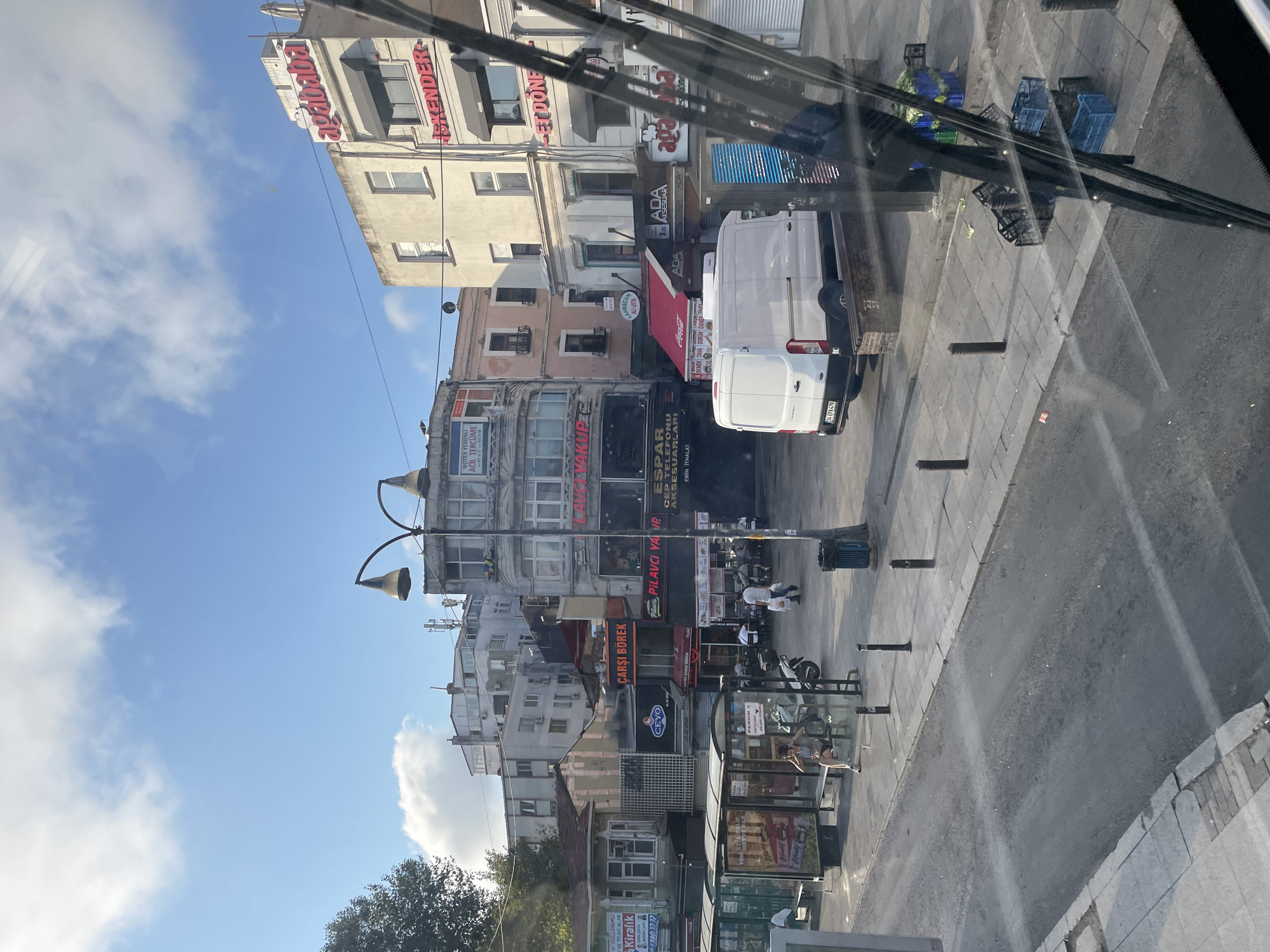 The image captures a street scene in Kadıköy, Turkey, on a sunny day with a partly cloudy sky. In the foreground, a bus windshield with wipers partially obscures the view of the street and buildings. The street itself appears to be paved with concrete and asphalt, with a sidewalk running alongside it.
Several buildings line the street, displaying a mix of architectural styles and commercial signage. Some of these signs are in Turkish, indicating businesses such as "AGABABA İSKENDER" and "PİLAVCI YAVUZ". A white van is parked on the street, and a bus stop shelter is visible on the sidewalk, with a woman sitting on a bench inside. Several street lamps and bollards are also present. The environment suggests a busy urban area with shops and pedestrian traffic.