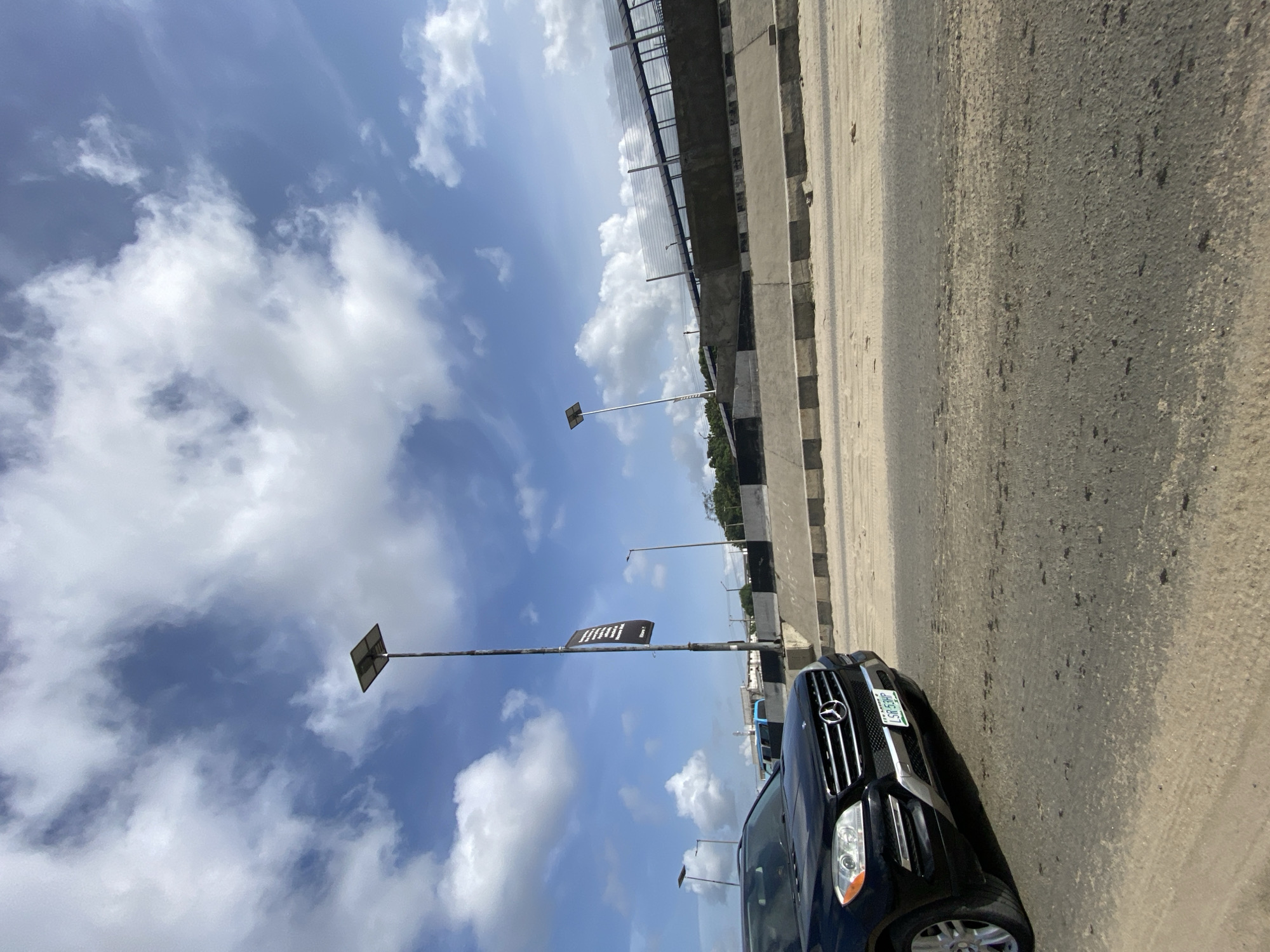 The image shows a black Mercedes-Benz SUV parked on the shoulder of a road next to a concrete barrier, possibly along a highway or bridge in Lekki, Nigeria. The license plate is visible. Overhead, a bridge spans the road. Blue sky with white clouds is overhead. Several streetlights line the road. The asphalt road shows signs of wear.