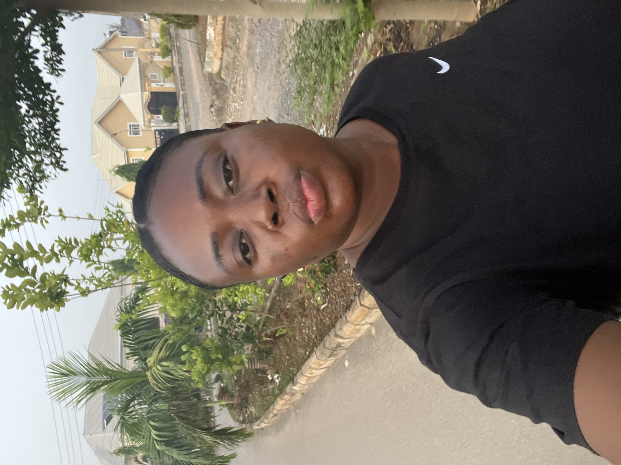 A person is taking a selfie outdoors on a street in Abuja, Nigeria. The person has dark skin and is wearing a black t-shirt with a white Nike swoosh logo on the right shoulder. The background includes a residential building with a beige exterior and a multi-peaked roof, lush green trees and shrubs, and a paved road. The weather appears to be overcast.