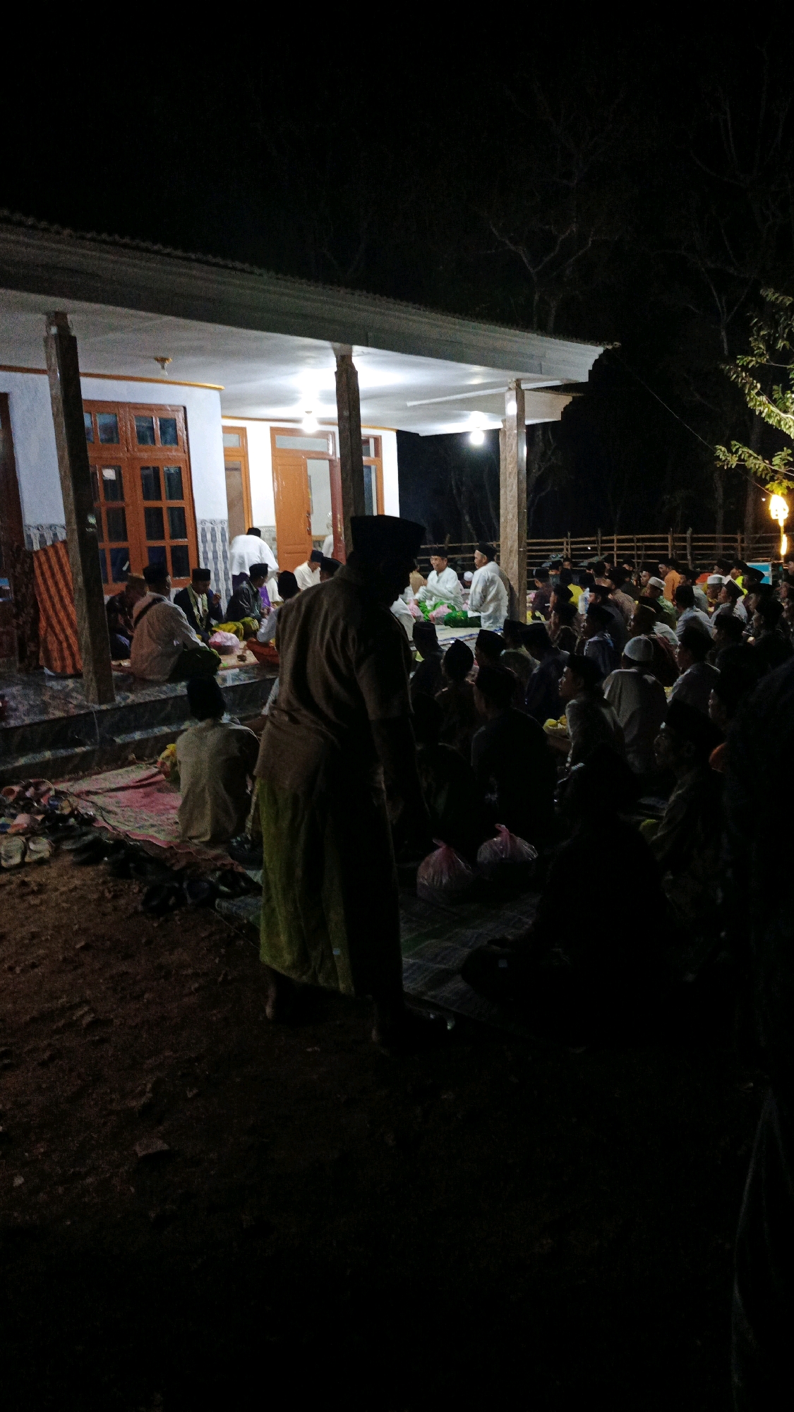 The image depicts an outdoor gathering at night in Sokaramme Timur, Indonesia, with a building in the background. Approximately fifty to seventy individuals, predominantly male, are seated on the ground on mats. Many are wearing traditional Islamic attire, including peci caps and sarongs, and some wear white robes. The lighting appears to be artificial, with bulbs illuminating the area around the building. The foreground is dark, with a few objects like bags visible. The ground is covered with dirt and scattered debris. The scene suggests a communal event, possibly a religious or social gathering, taking place after sunset.