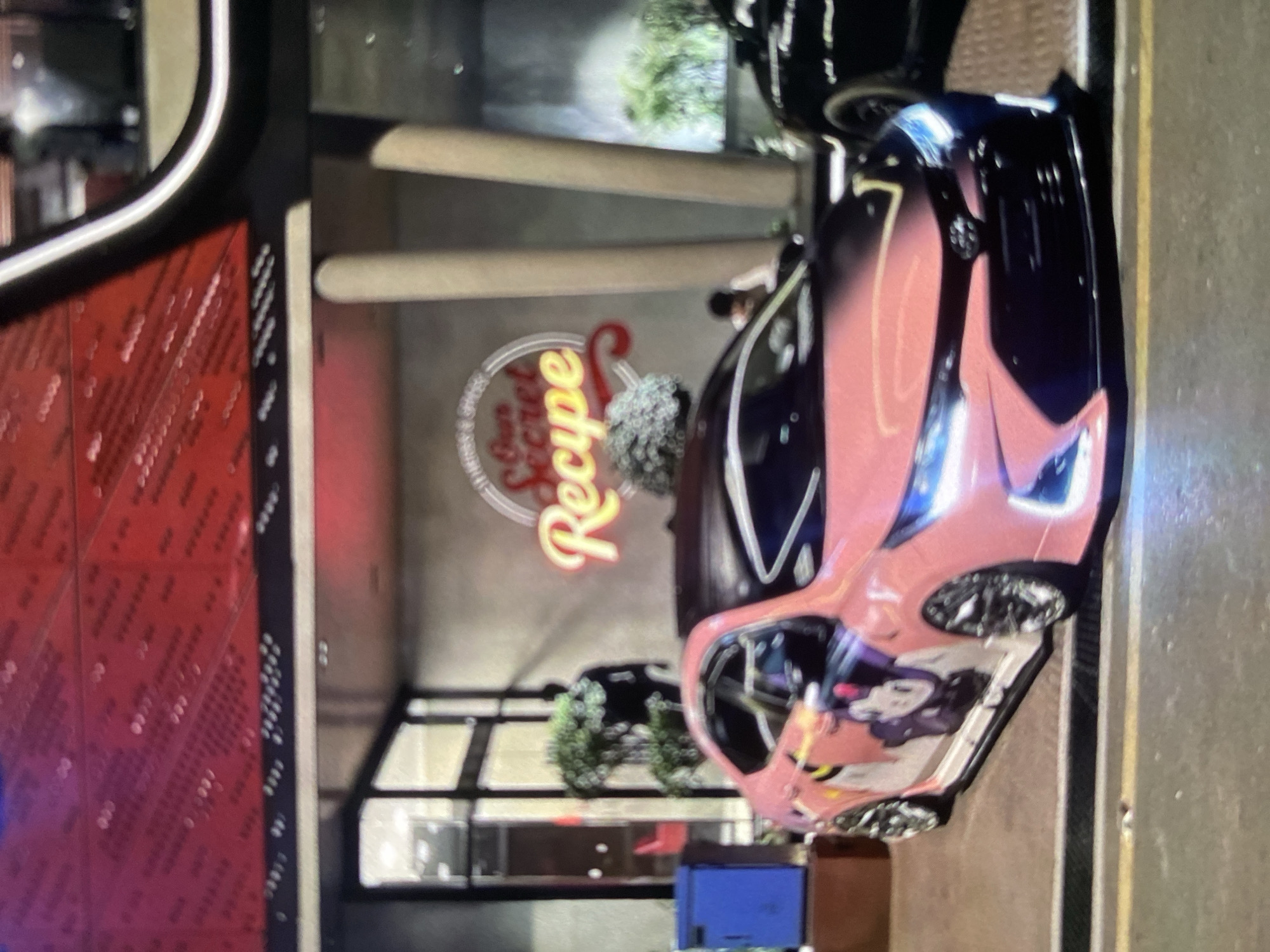 The image depicts a pink and black car parked on a street near a building with a neon sign reading "Our Secret Recipe." The location appears to be Lhokseumawe, Indonesia, based on the signage and general environment. There is a second car behind the first. The overall setting suggests a commercial or urban area. Other objects visible include a blue trash bin and some potted plants. The image appears to be taken at night.