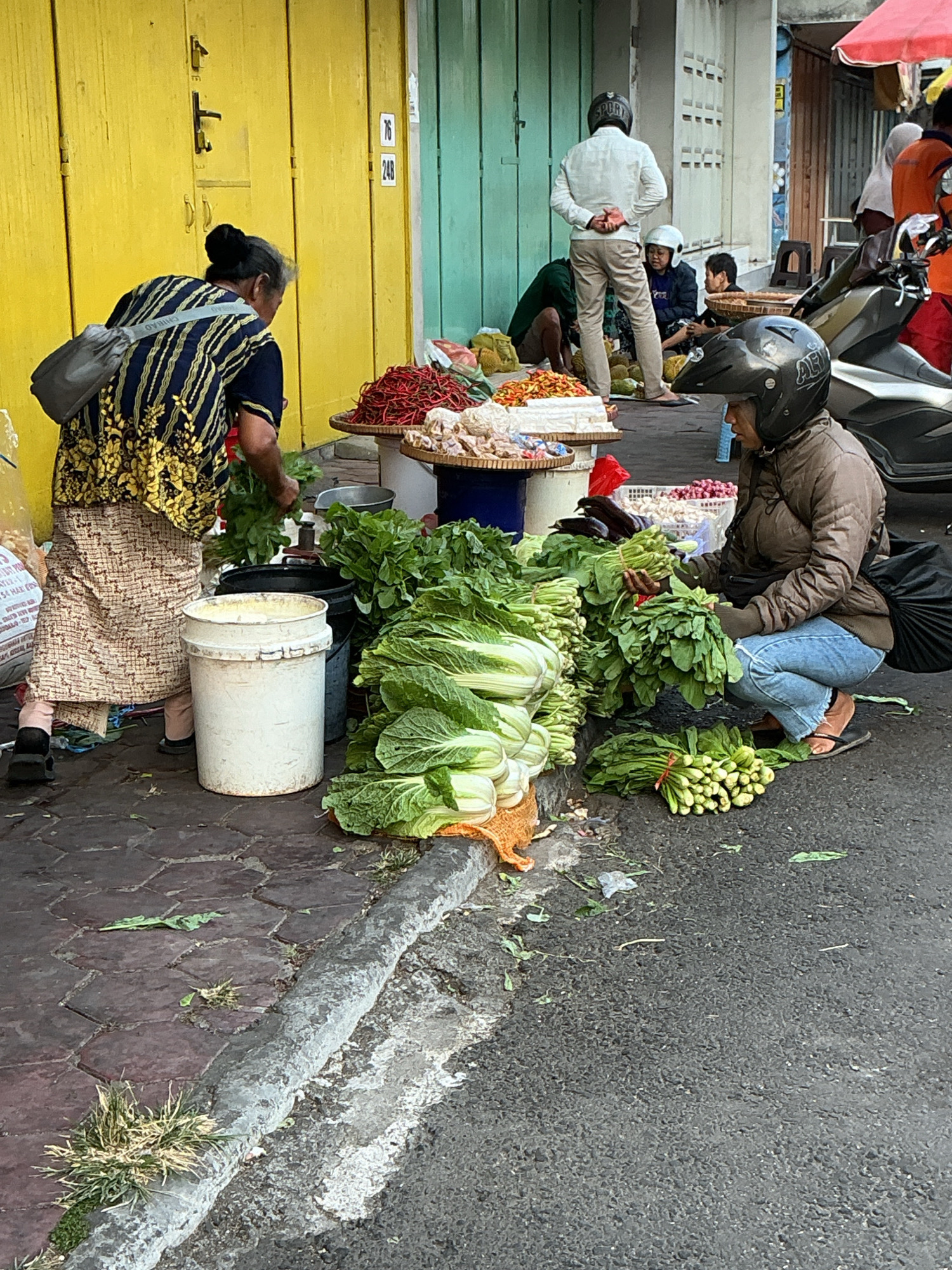 The image depicts a street market scene in Magelang, Indonesia. In the foreground, a woman wearing a helmet and a brown jacket is crouching down, holding a bunch of green leafy vegetables. She is surrounded by piles of fresh produce, including what appears to be napa cabbage and other leafy greens. To the left, another woman, older and wearing a patterned top and skirt, is attending to vegetables. Behind them, a yellow building with green doors serves as a backdrop. Several other individuals are visible in the midground and background, some wearing helmets, suggesting a motorcycle-friendly environment. The ground is a mix of paved stones and asphalt, with some debris scattered around. The overall impression is a busy, open-air market with vendors and customers interacting amongst fresh produce.