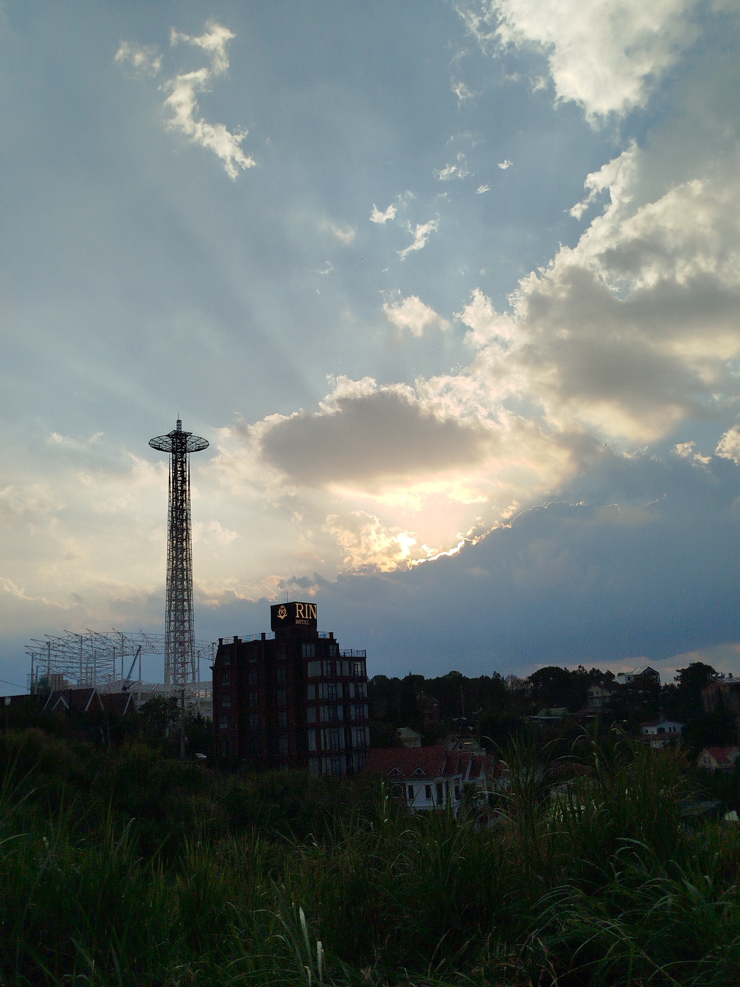 The image depicts a cityscape under a cloudy sky, likely in Da Lat, Vietnam. In the middle ground, there is a tall observation tower and a hotel building with the name "RIN HOTEL" prominently displayed on top. The tower is a lattice structure with a disc-shaped platform at the top. Adjacent to it, scaffolding indicates ongoing construction. The foreground is filled with tall grass and foliage. In the background, the silhouette of hills dotted with houses and trees are visible. The sun is partially obscured by the clouds, with rays of light streaming through.