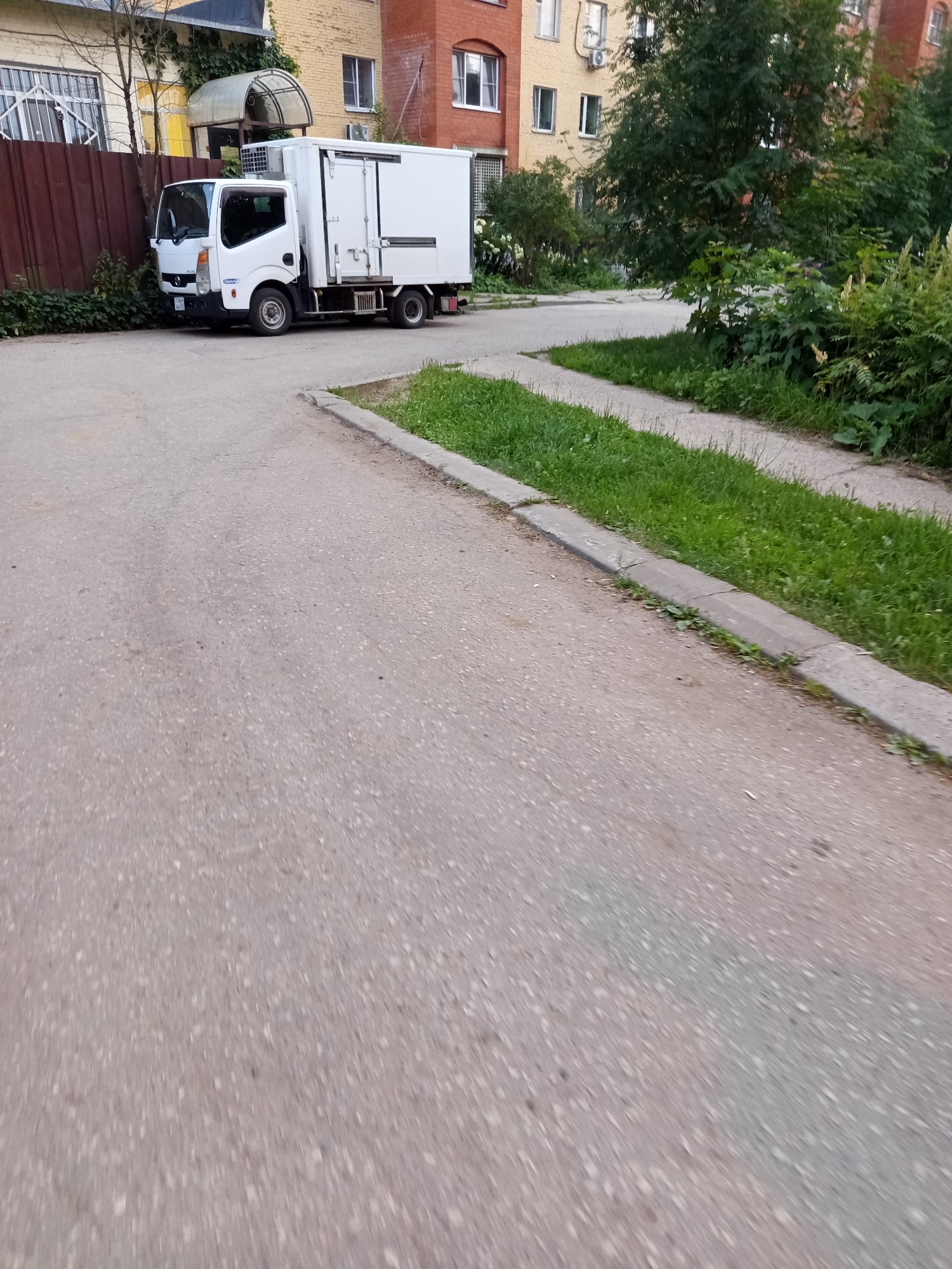 The image shows a white refrigerated truck parked on a paved area next to a building in Selyatino, Russia. The truck is facing left and has its rear doors closed. The building is multi-story with brick and yellow facade sections. A brown fence is visible to the left of the truck, and a sidewalk with green grass runs alongside the paved area. Trees and bushes are present around the building and the sidewalk. The sky is not visible in the frame.