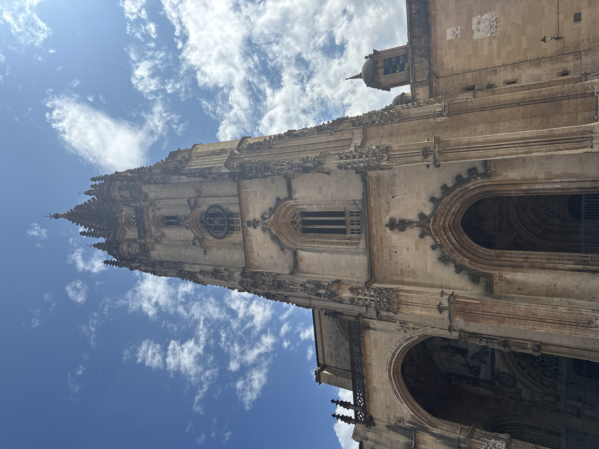 The image shows a low-angle view of the Oviedo Cathedral in Oviedo, Spain, on a day with a blue sky and scattered clouds. The cathedral's facade is made of light-colored stone, with intricate Gothic architectural details. The main tower, a prominent feature, is tall and slender, adorned with spires and ornamental carvings. A clock face is visible on the tower. Below the tower, there are large arched entrances to the cathedral, one of which is in deep shadow. The building exhibits a weathered appearance, typical of historic architecture. To the right of the main facade, a smaller bell tower with a domed roof is visible, as are portions of the cathedral's side walls with decorative stone carvings and small windows. The overall impression is of a grand and ancient religious structure bathed in sunlight.