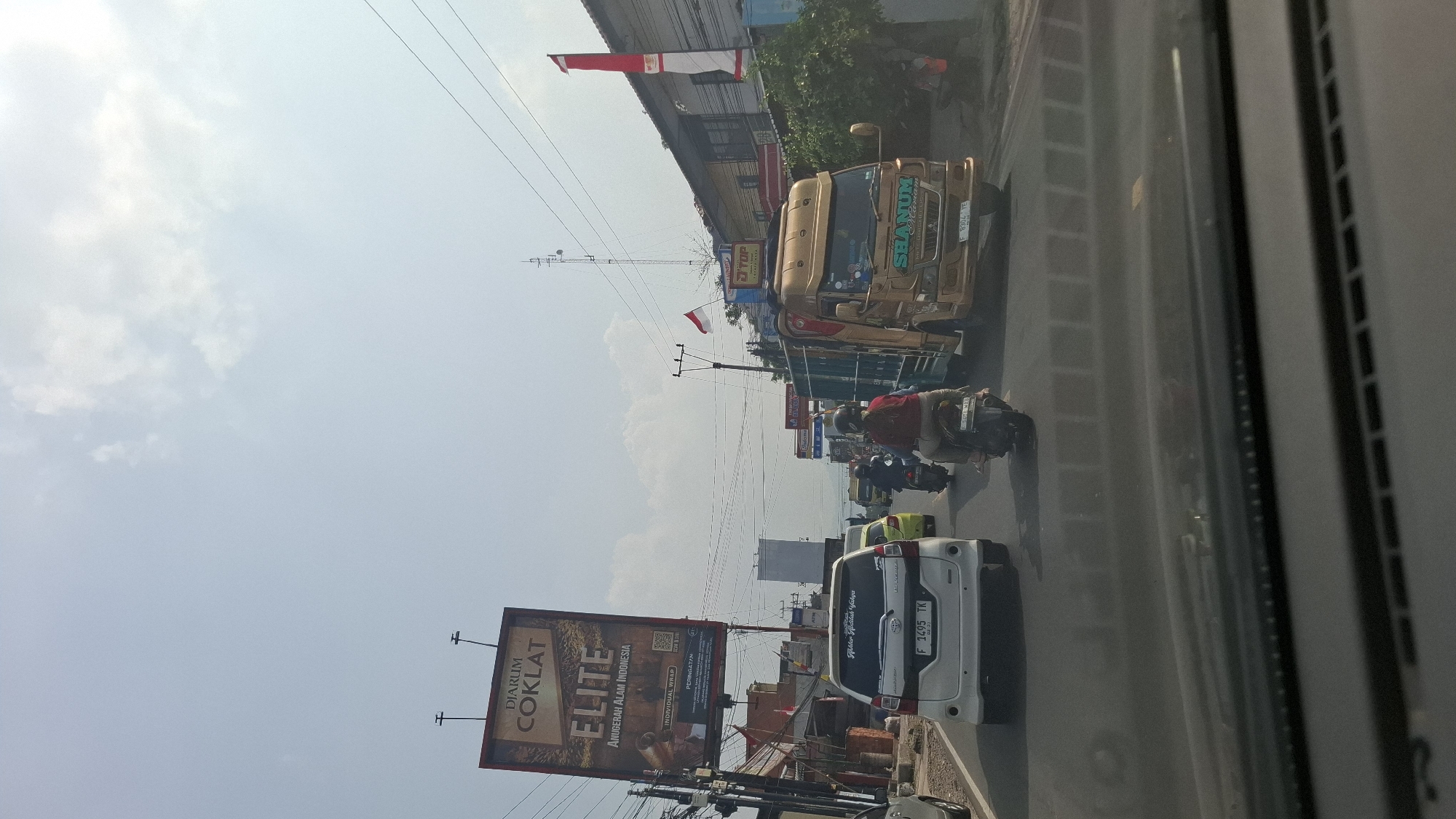 This image captures a street scene in Cikaroya, Indonesia, likely during daylight hours given the bright sky and clear visibility. The perspective is from inside a vehicle, looking forward.
The foreground is dominated by the dashboard and windshield of the vehicle from which the photo is taken. Beyond the windshield, a busy road is visible.
In the middle ground, several vehicles are in motion or stopped. On the left, a white Toyota Avanza with license plate "F 1495 TK" is prominent. To its right, a smaller yellow car is partially visible. Several motorcycles are also present, with one rider wearing a red shirt and another wearing a helmet.
A large, gold-colored truck is a significant element in the right half of the image. It has "SHANUM MELANIAM" written on its side. Behind this truck, buildings with awnities and signs can be seen. A tall telecommunications tower is also visible in the background.
Two Indonesian flags, red and white, are displayed on poles, one on a building and another on a utility pole, suggesting a national holiday or a significant event.
A large billboard for "DJARUM COKLAT ELITE" advertising cigarettes is positioned on the left side of the road, above the street level. The billboard features imagery related to chocolate and the tagline "ANUGERAH ALAM INDONESIA".
The environment appears to be a typical urban or semi-urban street in Indonesia, with overhead wires and utility poles lining the road. The sky is partly cloudy.