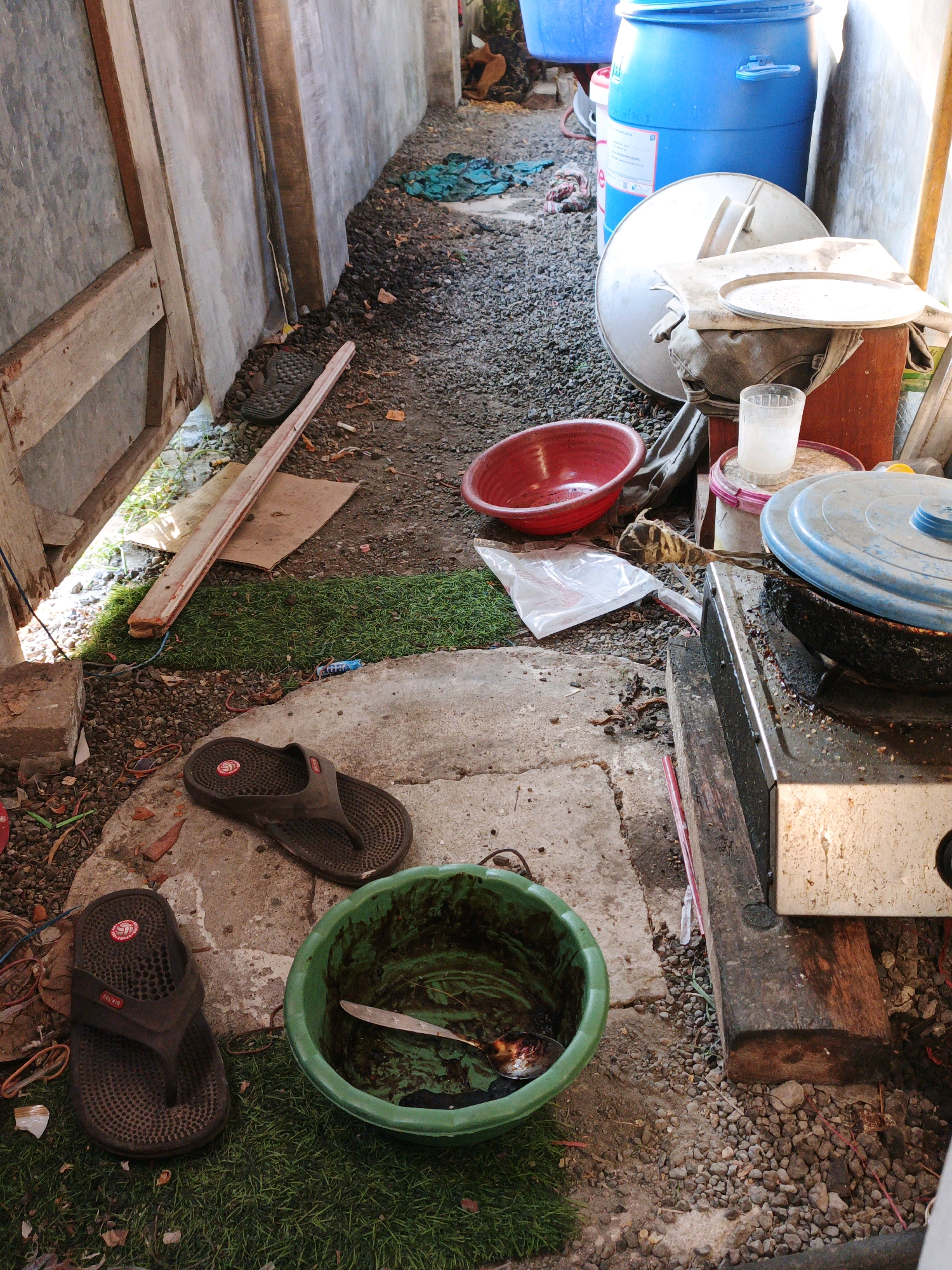 The image depicts a cluttered outdoor area, possibly a backyard or alleyway, in Ketegan, Indonesia. The ground is covered in gravel and concrete. Several objects are scattered around, including a large blue plastic barrel, a metal pot with a blue lid, a red bowl, a glass of water, a dirty green bowl containing a knife and what appears to be food scraps, and a pair of dark-colored flip-flops. A propane stove or similar cooking apparatus is visible, placed on wooden planks. There is also some green artificial turf laid out on the ground, and construction materials like wood planks and bricks are present. The scene suggests a lived-in, possibly unkempt, domestic environment. No people are visible in the image. The lighting suggests it is daytime.