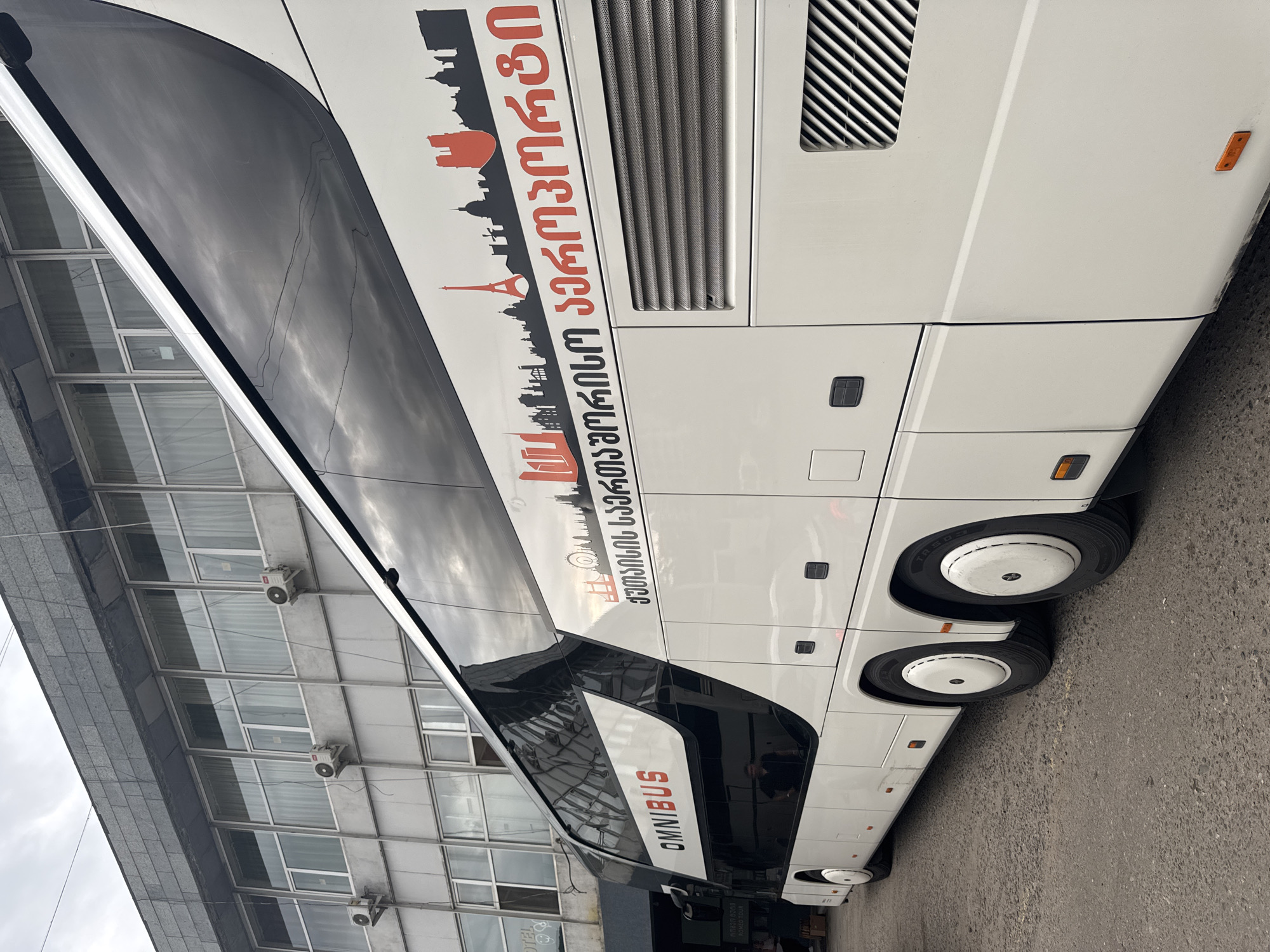 The image shows the side of a white tour bus with a black and red graphic depicting famous landmarks. The graphic includes silhouettes of the Eiffel Tower and the Arc de Triomphe, suggesting a European theme. The bus has the word "OMNIBUS" printed in white letters on a black background along its lower section. The bus is parked on a paved surface, possibly a street or parking lot, with a modern building featuring large windows in the background. The building's facade is reflective, mirroring the sky and clouds, which appear overcast, indicating a cloudy or overcast day. The overall scene suggests a stop during a tour or at a bus station in Tbilisi, Georgia. There is no visible text on the building or immediate surroundings, apart from the text on the bus itself. The image is taken from a slightly low angle, looking up at the bus.