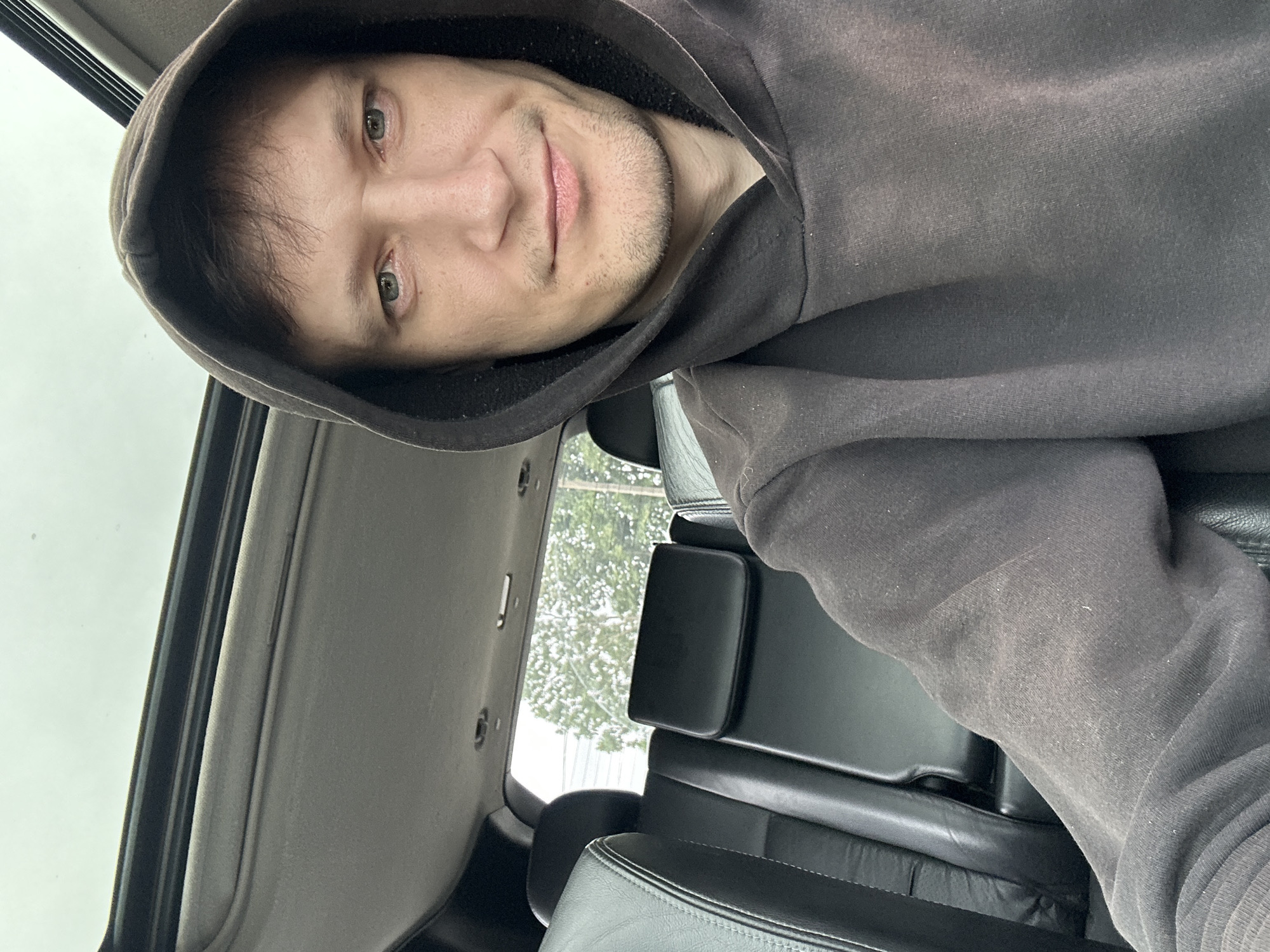 The image is a selfie taken from inside a car. The subject, a man wearing a dark hoodie, is looking at the camera. He has light skin, light-colored eyes, and stubble. The background shows the interior of the car, including black leather seats and a sunroof. Outside the car, trees and a cloudy sky are visible. The location is stated to be Makiv, Ukraine, but there are no visual cues in the image to confirm this.