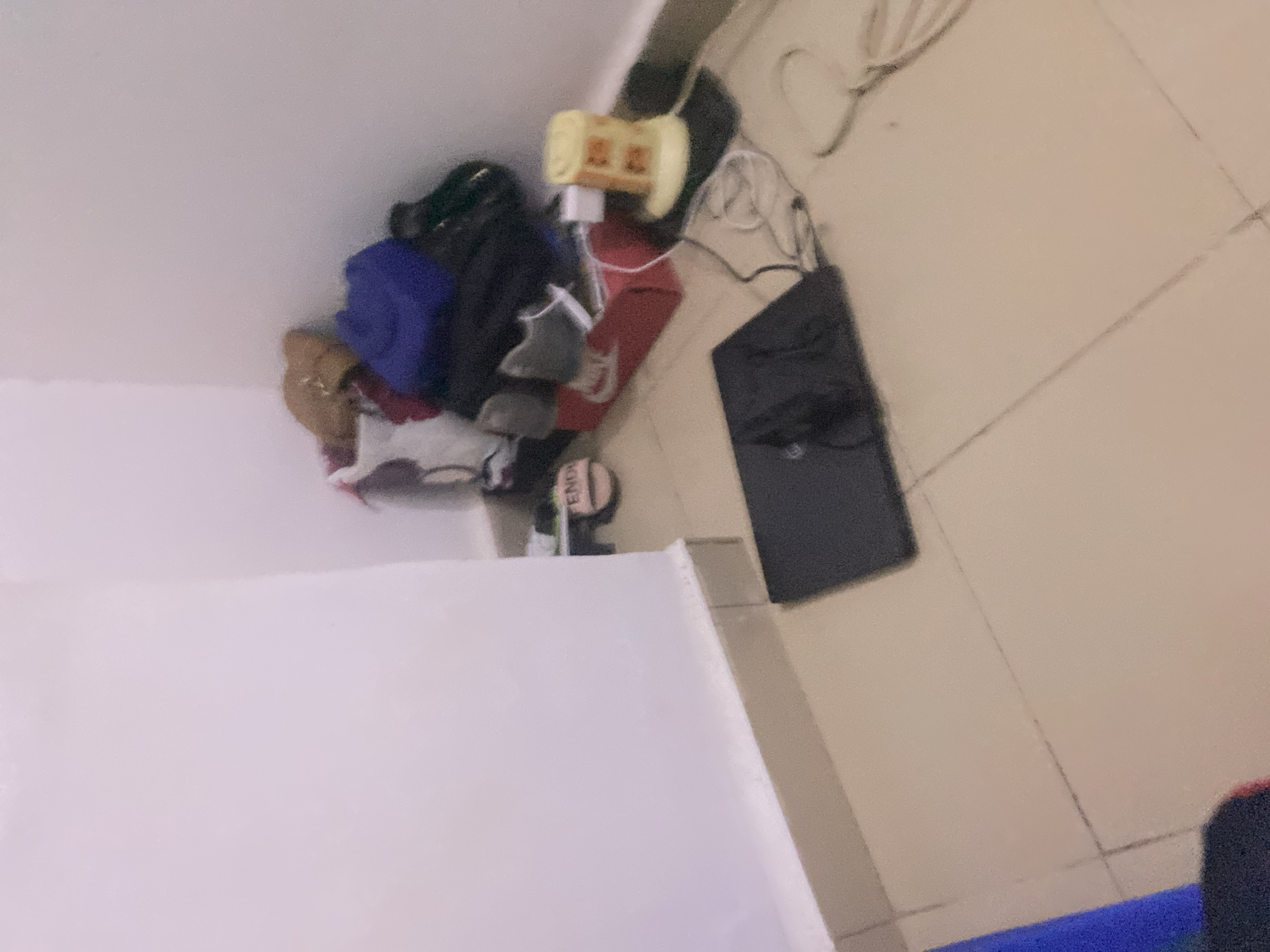 The image depicts a cluttered corner of a room. A pile of clothing and shoes is visible against a white wall. Among the items are a blue garment, a brown scarf or hat, and a pair of pink sandals with the word "FENDI" partially visible. Below the pile, a red shoebox with a Nike logo is present, and a grey shoe is resting on top of it. To the right of the shoebox, a yellow electric fan or heater is on the floor. A black laptop is open on the floor in the foreground, with cables connected to it. The floor is tiled in a neutral color. There are no people visible in the image. The scene appears to be indoors, in a residential setting, and it is not possible to determine the specific location in Howayi, Nigeria based solely on the visual information provided.