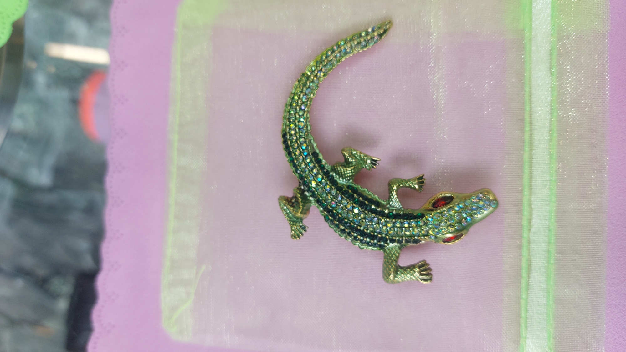 A close-up photograph features a decorative, gold-colored alligator brooch or pin, covered with sparkling stones. The alligator is placed on a pink and semi-transparent white fabric. Part of a purple surface can be seen to the left of the fabric. The setting and location are not discernible from the image.