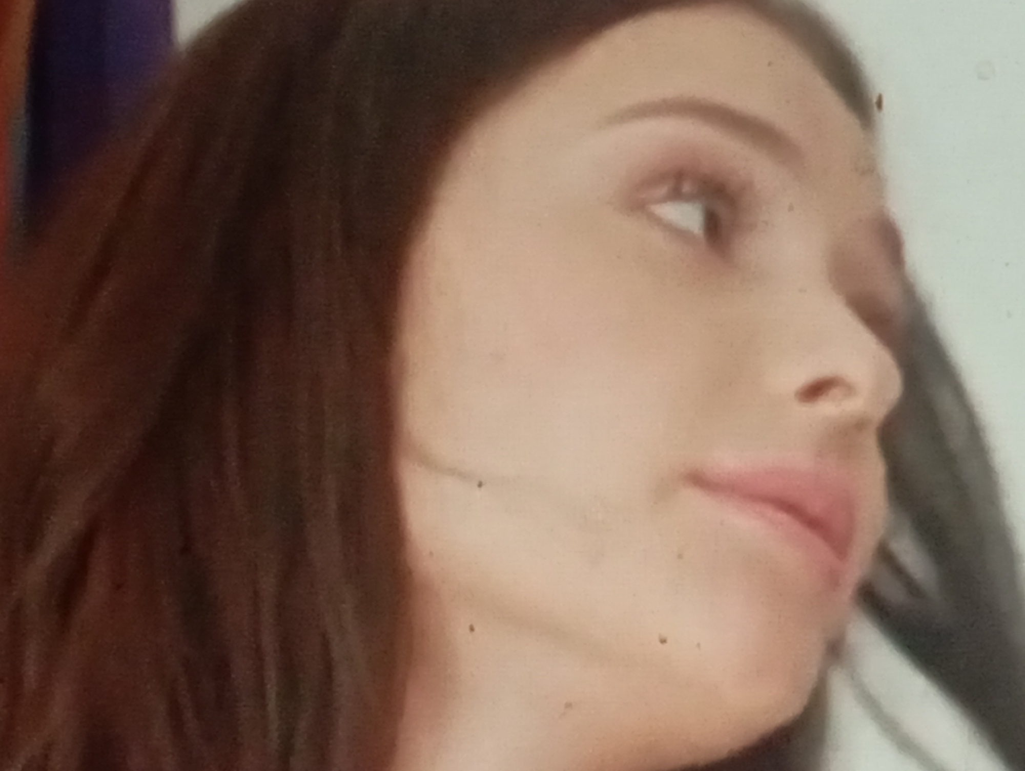 The image is a close-up, side profile of a young woman's face, with her head tilted slightly upwards. Her hair is dark brown and cascades down her left side. Her skin is fair with some scattered freckles. Her right eye is visible, appearing light brown or hazel with a subtle hint of mascara. Her lips are slightly parted and appear to be a natural pink hue, possibly with a light lip gloss. The lighting suggests an indoor setting, with a soft, diffused light source illuminating her face. The background is blurred, with hints of dark blue and a light, neutral color, making it difficult to ascertain the specific environment. There is no indication of any specific location within Sofiivska Borshchahivka, Ukraine, as the image focuses solely on the woman's facial features and does not contain any recognizable landmarks or contextual elements of that city.