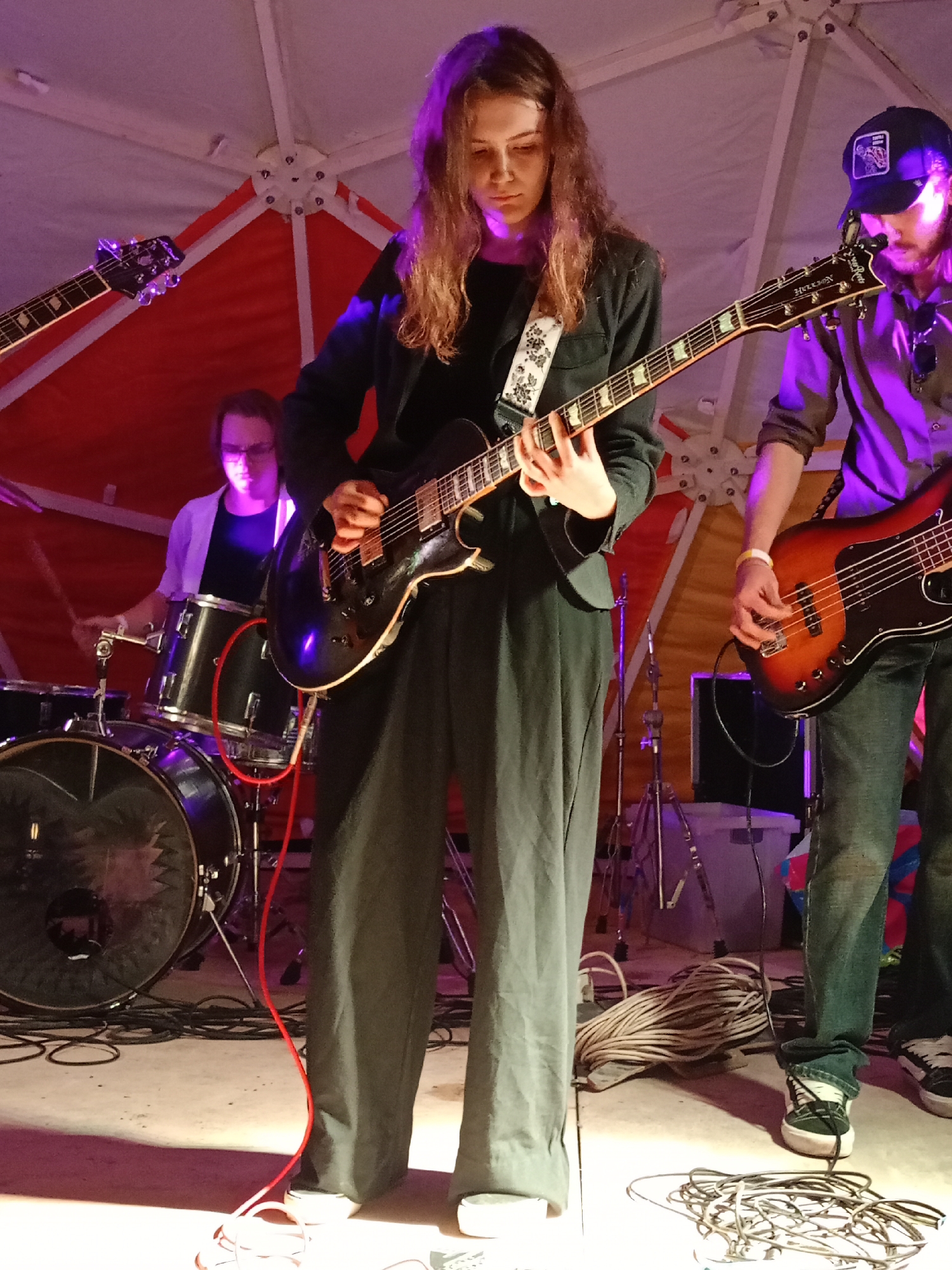 A band performs on stage in what appears to be a geodesic dome structure, with red fabric lining the exterior and a white interior. The lighting is a mix of purple and pink hues. On the left, a drummer, wearing glasses and a light-colored open shirt over a dark t-shirt, plays a drum set. In the center, a young woman with long, wavy brown hair, dressed in a dark jacket and dark pants, plays an electric guitar. She wears a strap with a white and black floral pattern. To the right, a person wearing a dark baseball cap and a dark shirt plays a bass guitar. They are wearing blue jeans and white sneakers with black accents. Several black cables snake across the floor, some of which are bundled up. The stage floor is concrete. The location is identified as Barnaul, Russia.