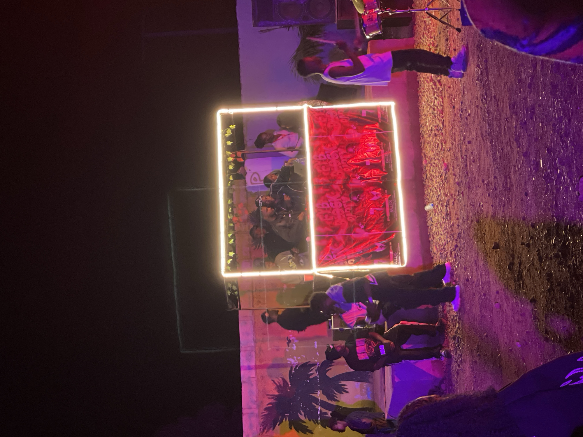 This image depicts an outdoor night scene in Durumi, Nigeria, characterized by vibrant purple and pink lighting. A group of individuals are gathered around a brightly illuminated, rectangular stage structure adorned with a red banner displaying what appears to be album art. A DJ setup is visible within the structure, with several people standing and interacting.
In the foreground to the left, a person with dark, curly hair is partially visible, facing away from the camera. To the right of the stage, a young man is playing drums, dressed in a white tank top and dark pants. Behind him, a large speaker system is positioned against a wall.
The background features a wall painted with a mural that includes palm trees. Several individuals are seated or standing on a raised platform in front of the painted wall. The ground is covered in a mix of gravel and dirt. The overall atmosphere suggests a live music event or gathering.