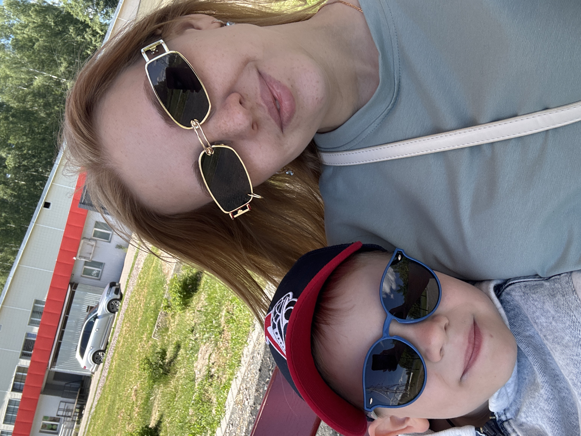 An outdoor, rotated image (90 degrees counter-clockwise) features two individuals, an adult woman and a young child, posing for a photo on a sunny day.
The adult, likely a woman, is positioned in the upper part of the frame. She has long, light brown hair, fair skin with visible freckles, and wears distinctive rectangular sunglasses with thick gold frames. Her expression is a soft smile. She is wearing a light blue-green top, and a white strap, possibly from a bag, is visible across her chest.
Below and slightly to the right of the adult is a young child, whose face is mostly obscured by large, blue-framed round sunglasses. The child is wearing a dark baseball cap with a red brim and a white emblem, and a denim jacket is visible on their upper body. A faint smile can be seen on the child's lips. Reflections in both pairs of sunglasses show blue sky with clouds and green foliage, indicating bright, clear weather.
The background includes a grassy area, trees, and a light-colored car (possibly an SUV) parked near a building. The building has white walls and red trim along its roofline and possibly around windows. A red bench is partially visible in the foreground, suggesting they are seated. The overall mood of the image is relaxed and cheerful. No discernible text is visible on any object in the image.