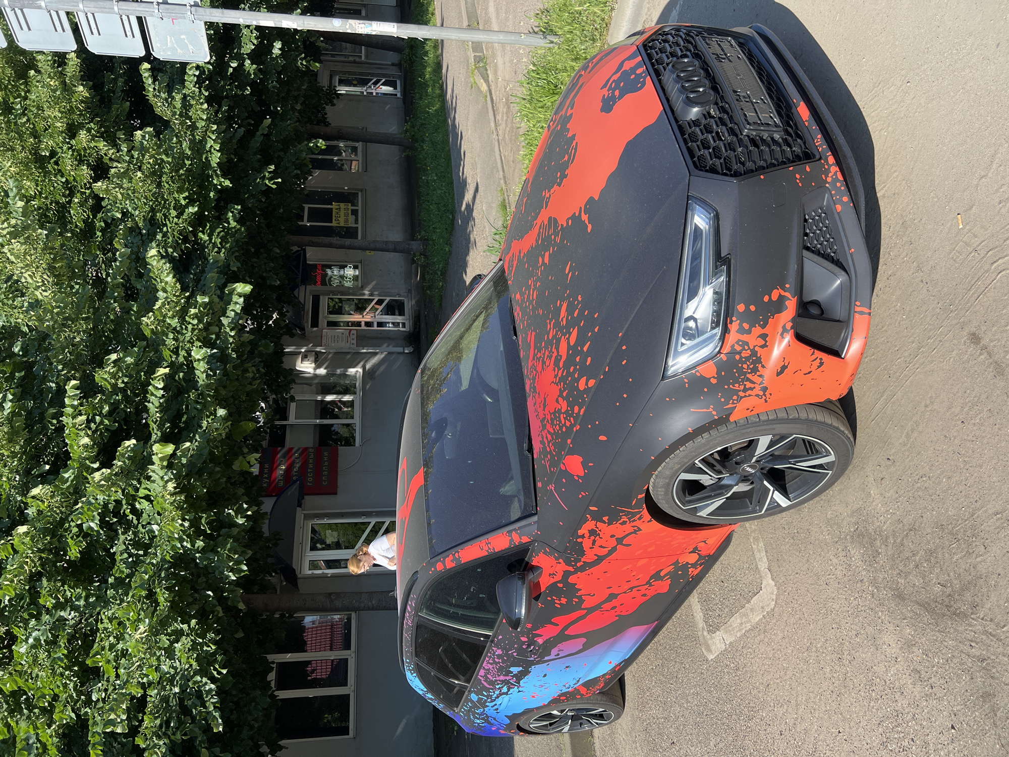 An Audi A4 B9 Avant (wagon), distinctively wrapped in matte black with a vibrant red, blue, and magenta paint splatter design, is parked on an asphalt surface under bright daylight. The custom wrap features red splatters predominantly on the hood and front fenders, transitioning to blue and magenta splatters along the side panels and rear. The vehicle is equipped with black and silver multi-spoke alloy wheels and a black honeycomb grille, characteristic of Audi's performance-oriented models. A license plate frame is visible on the front bumper, but no license plate is present or legible. Dust is evident on the windshield.
In the background, a light-grey commercial building with multiple windows and glass doors is visible. Cyrillic signage on the building identifies it as a furniture store, listing categories such as "КУХНИ" (KITCHENS), "ШКАФЫ" (WARDROBES), "ДЕТСКИЕ" (CHILDREN'S), "ГОСТИНЫЕ" (LIVING ROOMS), and "СПАЛЬНИ" (BEDROOMS). Another sign clearly displays "АРЕНДА" (RENT) with a phone number beginning 8-861, confirming the location as Krasnodar, Russia. Mature trees with dense green foliage line the front of the building, casting shadows across the scene.
A light-skinned female with light brown hair, tied back and wearing a white short-sleeved top, stands near one of the building's entrances, looking down. The parking surface is asphalt, exhibiting some wear, and features a faded, light-colored rectangular outline. A concrete curb separates the parking area from a strip of grass and weeds, which extends to a sidewalk. On the right side of the image, a metal pole with two stacked grey rectangular sign panels, likely traffic signs, stands adjacent to the road. A small piece of litter, possibly a cigarette butt, is visible on the asphalt in front of the car's bumper. The sunlight suggests a clear sky, with strong shadows indicative of midday or early afternoon.
