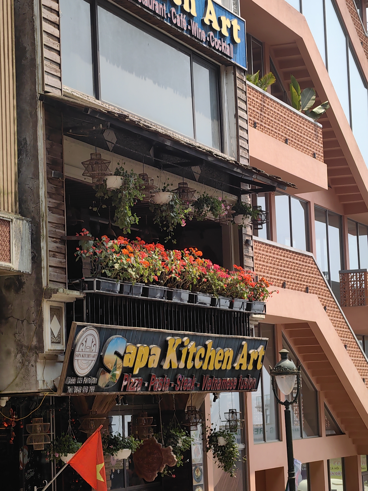 The image displays a partial view of two adjacent commercial buildings located on 029 Fansipan Street in Sa Pa, Vietnam, under clear daylight conditions.
The left structure, identified by a prominent sign as "Sapa Kitchen Art," is a multi-story building featuring a textured, weathered grey-brown facade with vertical wooden paneling. An upper-level sign, partially visible, indicates "Restaurant - Café - Wine - Cocktail." Below this, a balcony or recessed outdoor area contains numerous black rectangular planters filled with red and orange flowers. Above these flowers, several woven pendant light fixtures and hanging green plants in white pots are suspended. The large "Sapa Kitchen Art" sign, with yellow lettering on a dark background, also specifies "Pizza - Pasta - Steak - Vietnamese fusion" and provides a telephone number. A red Vietnamese flag with a yellow star is visible flying at an angle in the lower-left foreground.
Immediately to the right, a more contemporary multi-story building with a light peach-colored exterior is visible. This building features extensive glass windows and an external staircase characterized by a patterned terracotta-colored railing. Green leafy plants are discernible on an upper-level balcony behind this patterned railing.
In the lower-right foreground, an ornate, dark-colored street lamp with a white globe is positioned. No people or specific actions are visible in the scene.