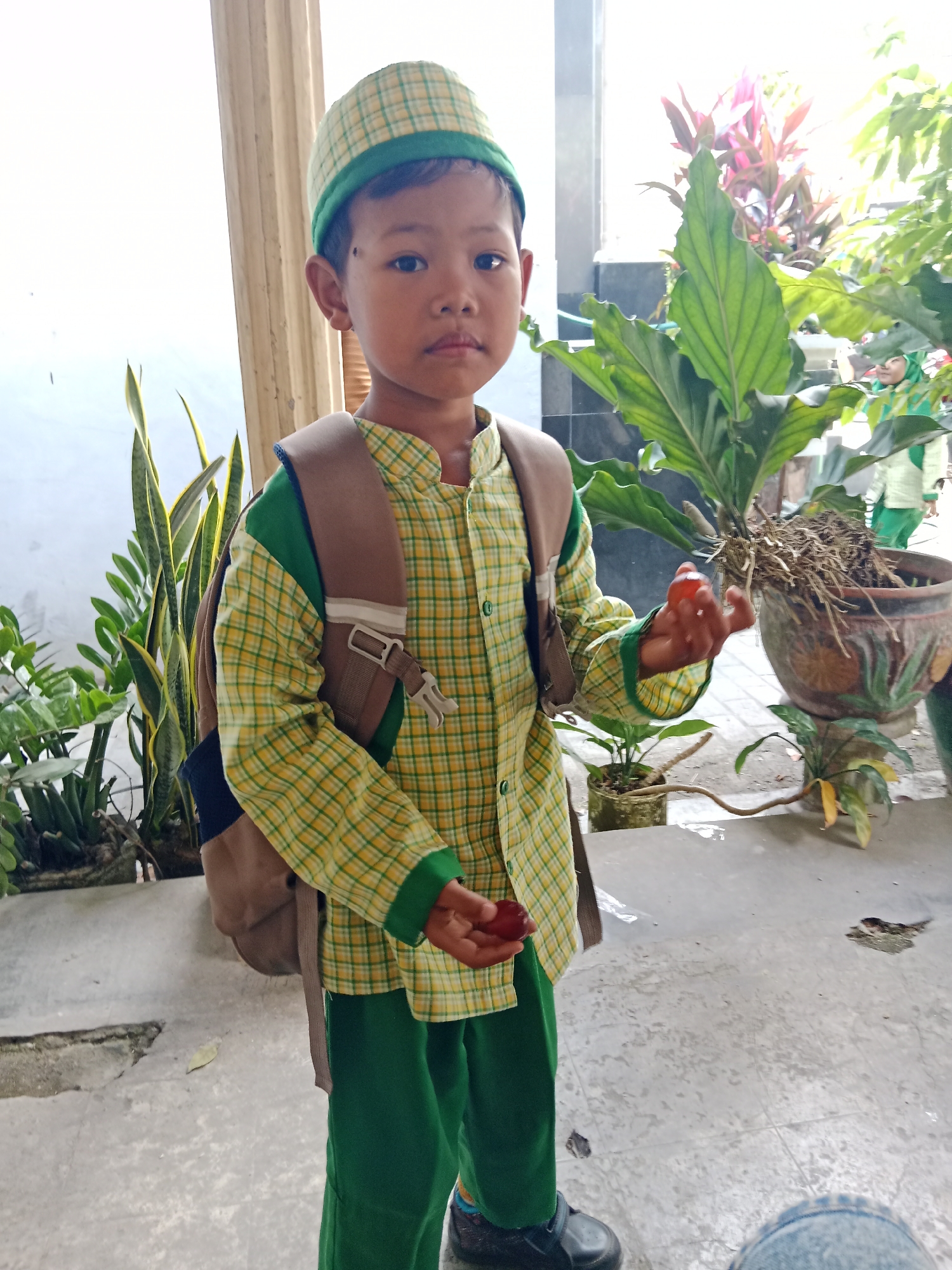 A young boy stands outdoors, facing the camera, in what appears to be the city of Srikaton, Indonesia. He is wearing a green and yellow checkered long-sleeved shirt with green trim and matching green pants. A brown backpack is slung over his shoulders. On his head, he wears a matching green and yellow checkered cap. In his right hand, he holds a small red object, and in his left hand, he holds another similar object. He is positioned in front of a large potted plant with broad green leaves. To his left, there are other plants, including a snake plant. The ground is paved with concrete. In the background, other people can be seen, including one wearing a green headscarf. The lighting suggests it is daytime.