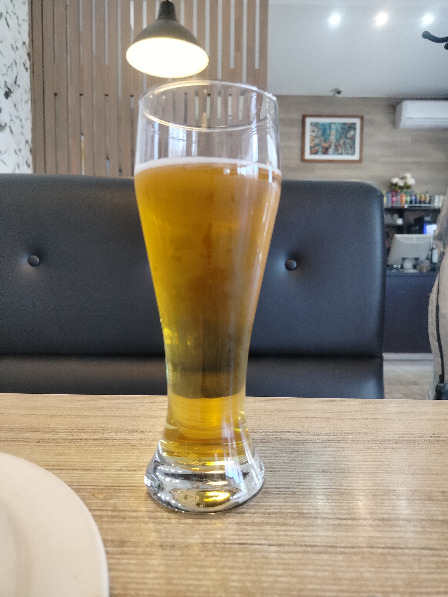 A glass of light beer sits on a light-colored wooden table in what appears to be a restaurant. Behind the glass is a dark gray booth with buttons. In the background, a painting hangs on the wall. There is a light fixture above the table and a white plate on the left side of the table, partially out of view. An air conditioner is mounted high on the wall in the background. The image was taken in Chaikovsky, Russia.