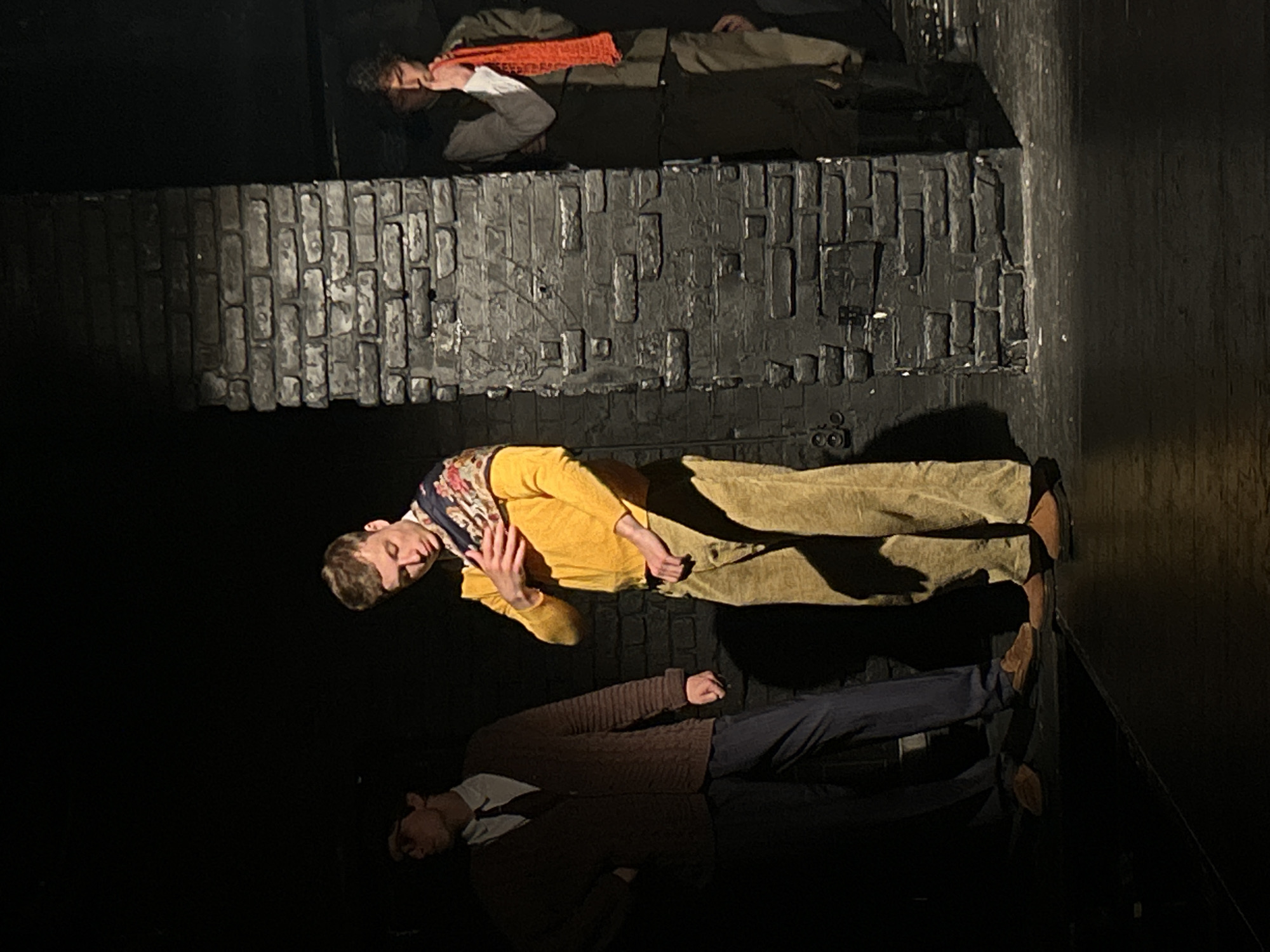 Three people lie on platforms of varying heights. The platforms are painted to resemble brick. The scene appears to be a stage production.