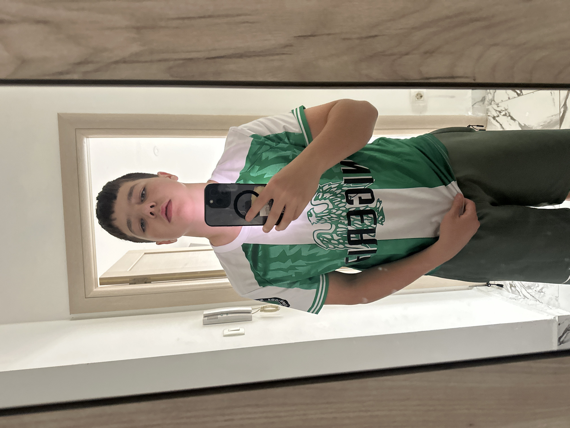A young male stands in front of a mirror, holding a black smartphone to take a selfie. He is wearing a green and white jersey with the word "MEXICO" printed on it, paired with green shorts. The background suggests an interior space, likely a bathroom, with a white countertop and a framed doorway visible in the mirror's reflection. Based on additional data (not in this image), the location is in Budva, Montenegro.