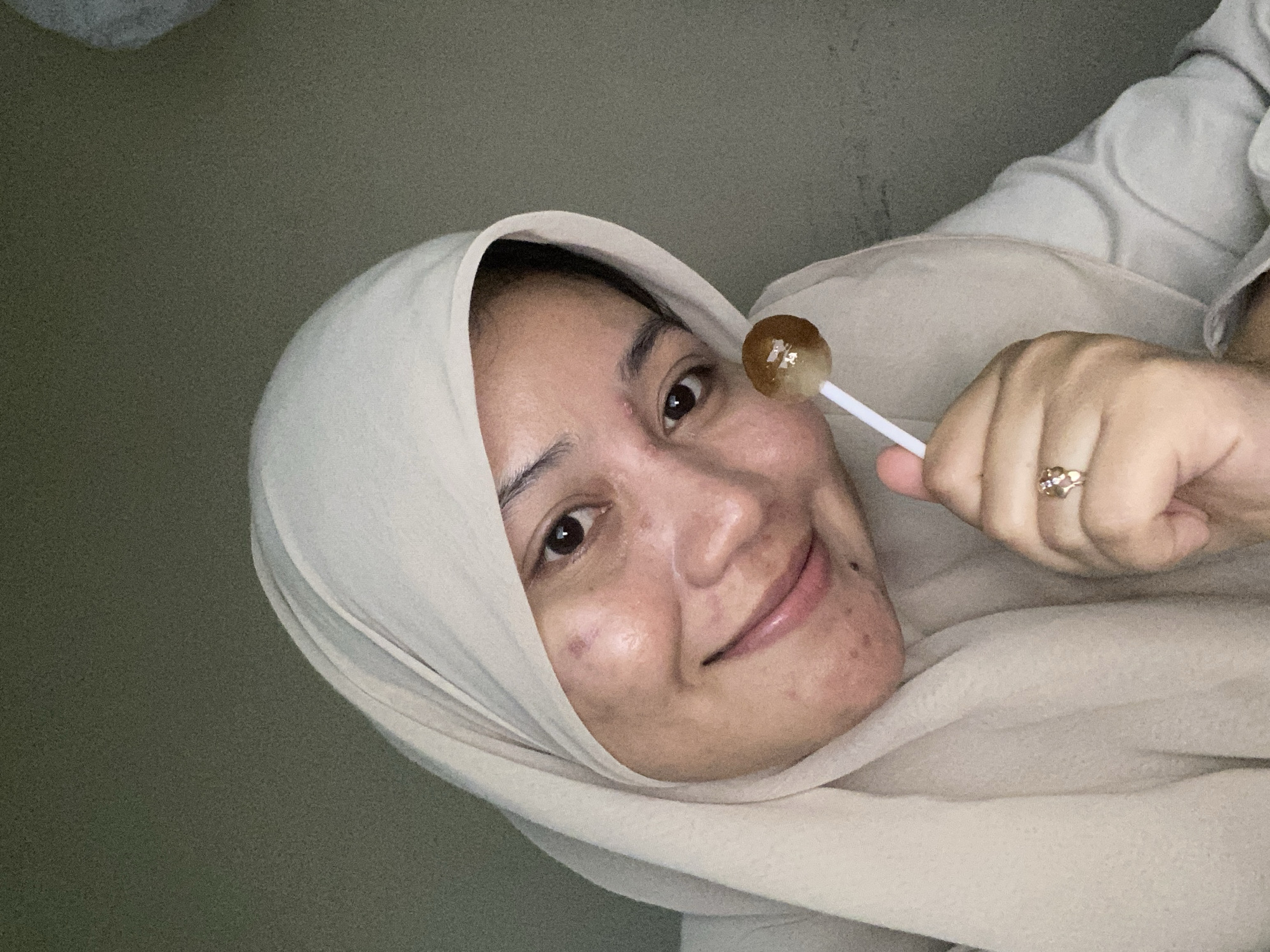 The media file shows a close-up, selfie-style shot of a young woman wearing a light-colored hijab. She is holding a lollipop to her cheek. The woman has a warm, smiling expression, and her eyes are looking towards the camera. The background appears to be an indoor setting with a plain, textured wall. The lighting suggests it is daytime, possibly indoors with artificial lighting. There is a ring on her finger. The overall mood of the image is casual and playful.