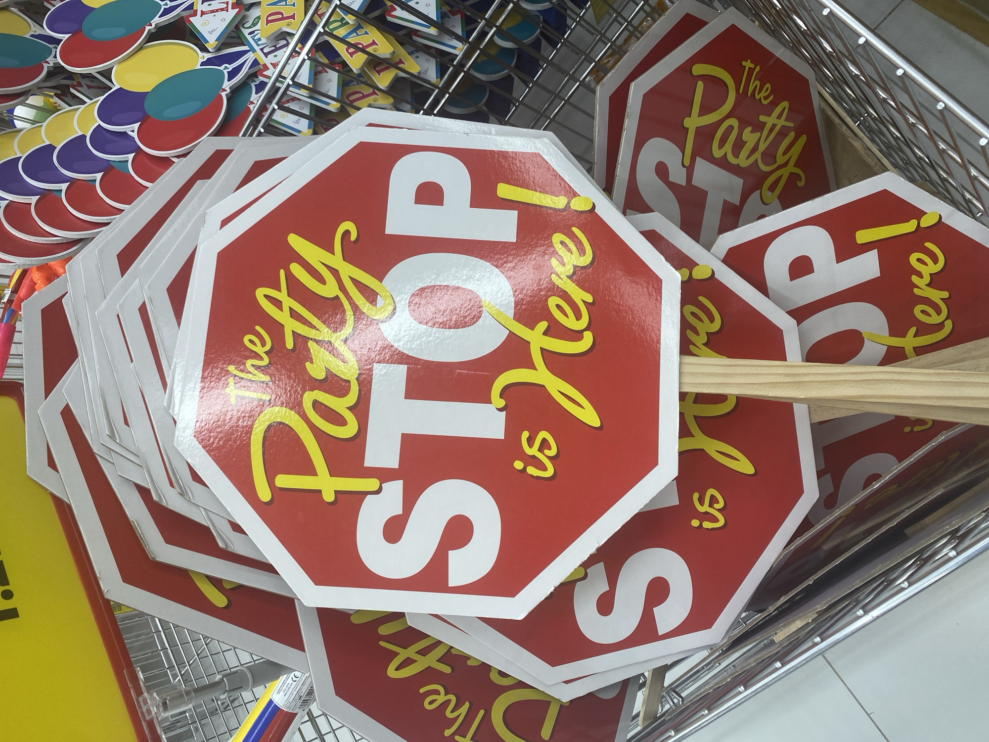The image shows a close-up of several red octagonal signs with white borders and yellow lettering. The signs read "The Party STOP is Here!". The signs are stacked in a metal wire basket, suggesting they are for sale in a retail environment. Some of the signs have wooden handles attached. In the background, other party decorations, including colorful balloons, are visible. The lighting is bright and appears to be indoor. There are no people in the image. The scene is a retail display of party decorations.