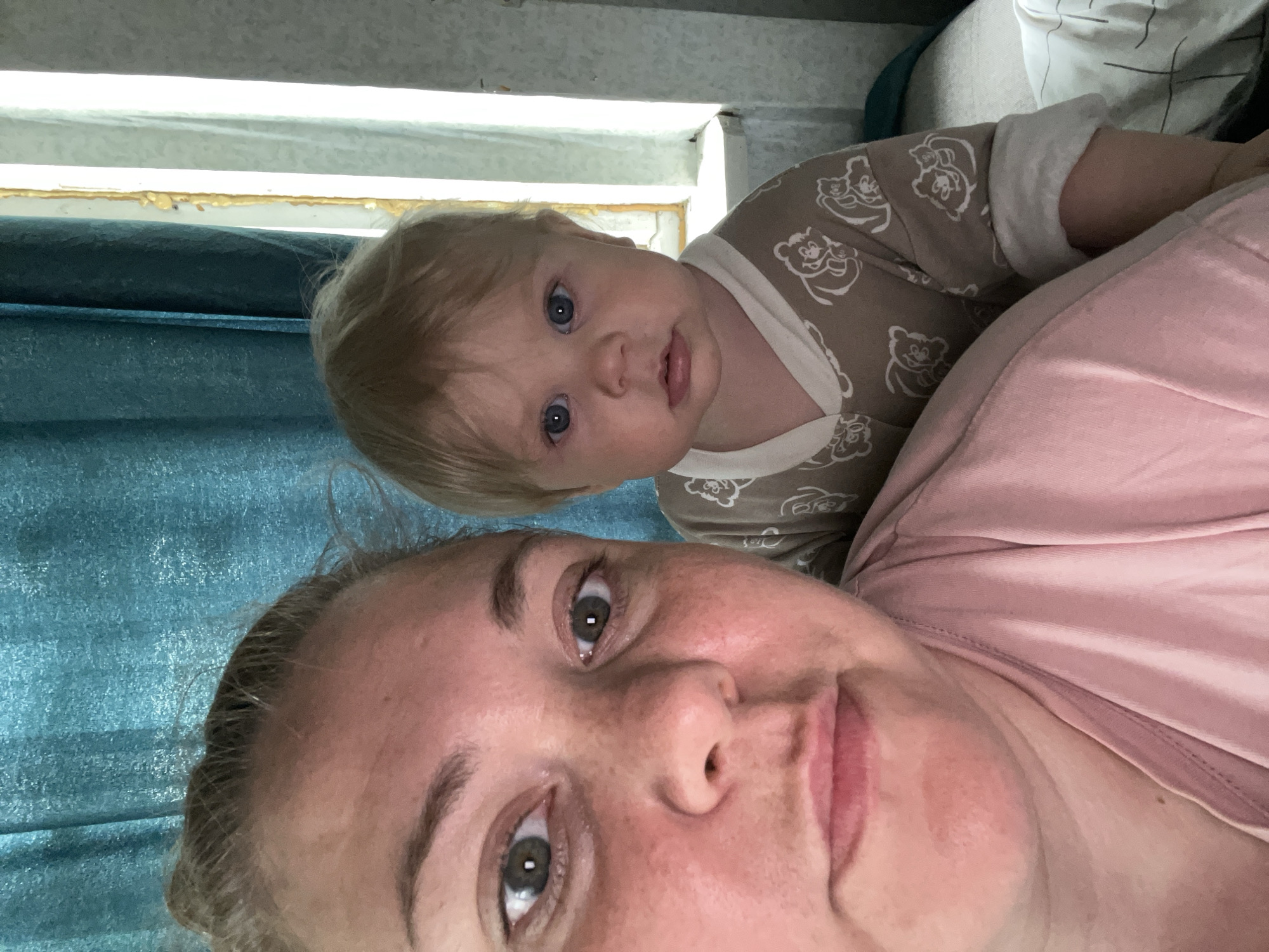 The image is a selfie featuring a woman and a baby. The woman, in the foreground, is looking directly at the camera with a neutral expression. She has fair skin, freckles, and her hair is pulled back. She is wearing a pink top. To her right and slightly behind her is a baby with blonde hair and blue eyes, also looking at the camera. The baby is wearing a light brown onesie with a pattern of bears. The background consists of a turquoise curtain on the left and a window with white blinds on the right. The setting appears to be indoors, likely a residential room. The image's metadata suggests it was taken in the city of Fokino, Russia.