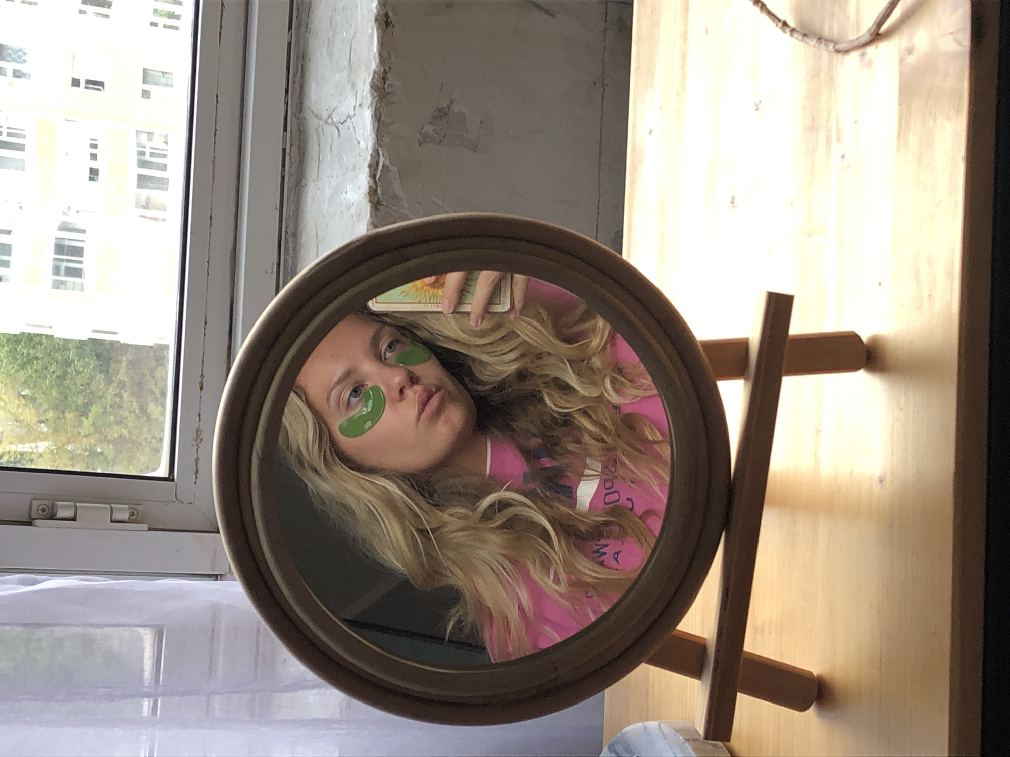 A young woman with long blonde hair and green under-eye masks is reflected in a round mirror. She is wearing a bright pink t-shirt. She holds a tarot card, the Sun, in her right hand. The mirror is on a wooden easel and rests on a light wooden table. In the background, through a window, a building and trees are visible. The setting appears to be an indoor space with a window overlooking an urban environment in Khimki, Russia.