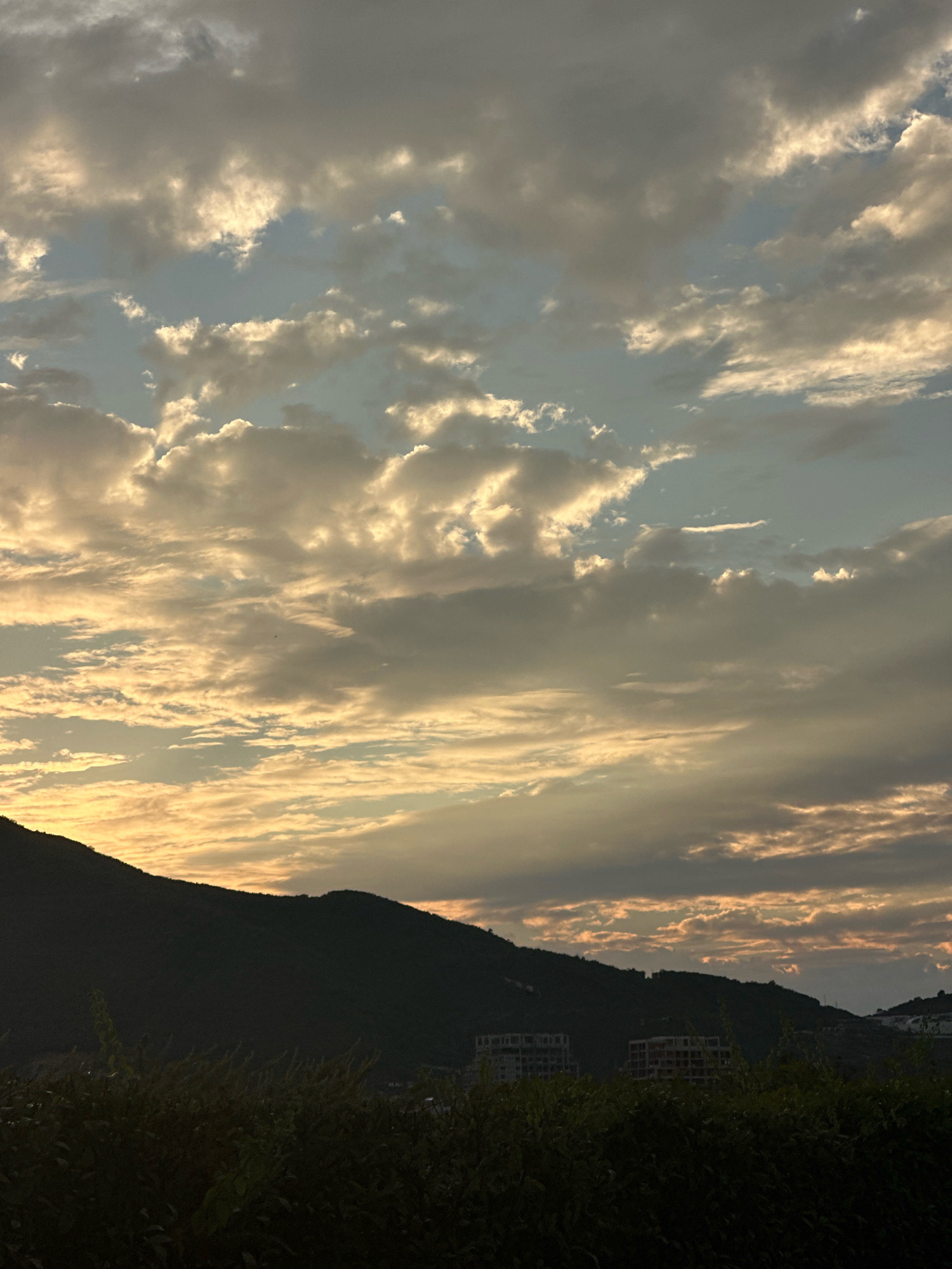 The image depicts a silhouetted mountain range against a sky filled with dramatic clouds during what appears to be either sunrise or sunset. The clouds are illuminated by the sun, creating a gradient of warm oranges and yellows, contrasted with cooler grays and blues. The lower portion of the image shows the dark silhouette of lush vegetation in the foreground, with hints of buildings visible on the slopes of the mountains in the mid-ground. These buildings appear to be under construction or are unfinished structures. The overall scene evokes a sense of natural beauty and the ongoing development of the landscape. This scene is likely located in Budva, Montenegro, a coastal town known for its mountainous backdrop and developing urban areas.