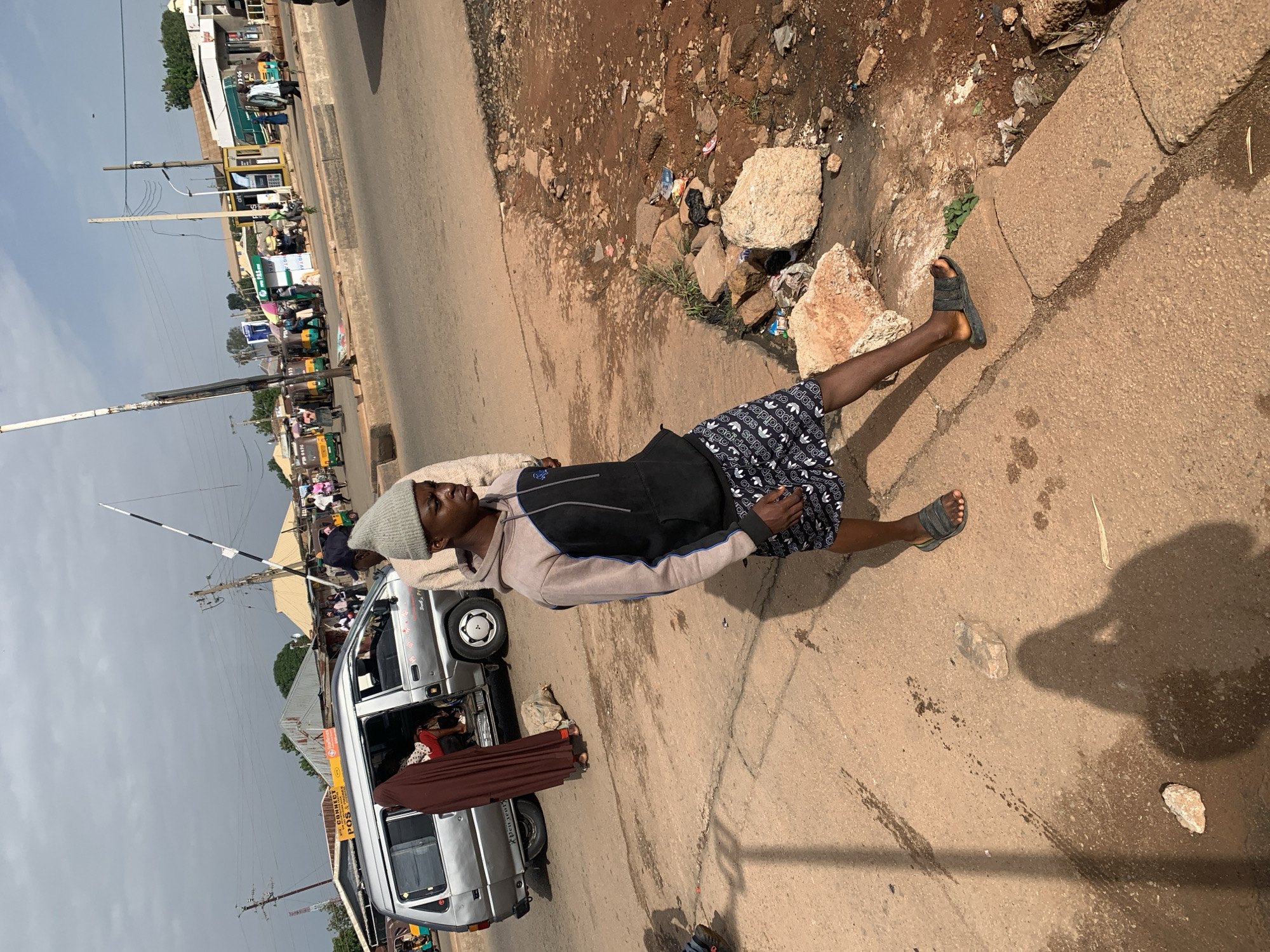 This is a medium shot, eye-level, outdoor, full-color photograph taken on a dusty street in Gura Top, Nigeria. The scene depicts a young man wearing a grey beanie, a light brown and black hoodie, and black patterned shorts, standing in the foreground, looking towards the left. He is wearing dark sandals and his right leg is extended forward. Behind him, a silver minivan is parked, with its side door open. A person covered in a dark brown hijab is standing near the open door of the minivan, partially obscuring the interior. In the background, there are several buildings, including shops and what appears to be a bus station with various colorful vehicles, including minibuses and tricycles. A traffic signal pole with a black and white striped barrier is visible. The street is paved with a reddish-brown dirt and asphalt, with some gravel and debris scattered along the roadside. The sky is partly cloudy.