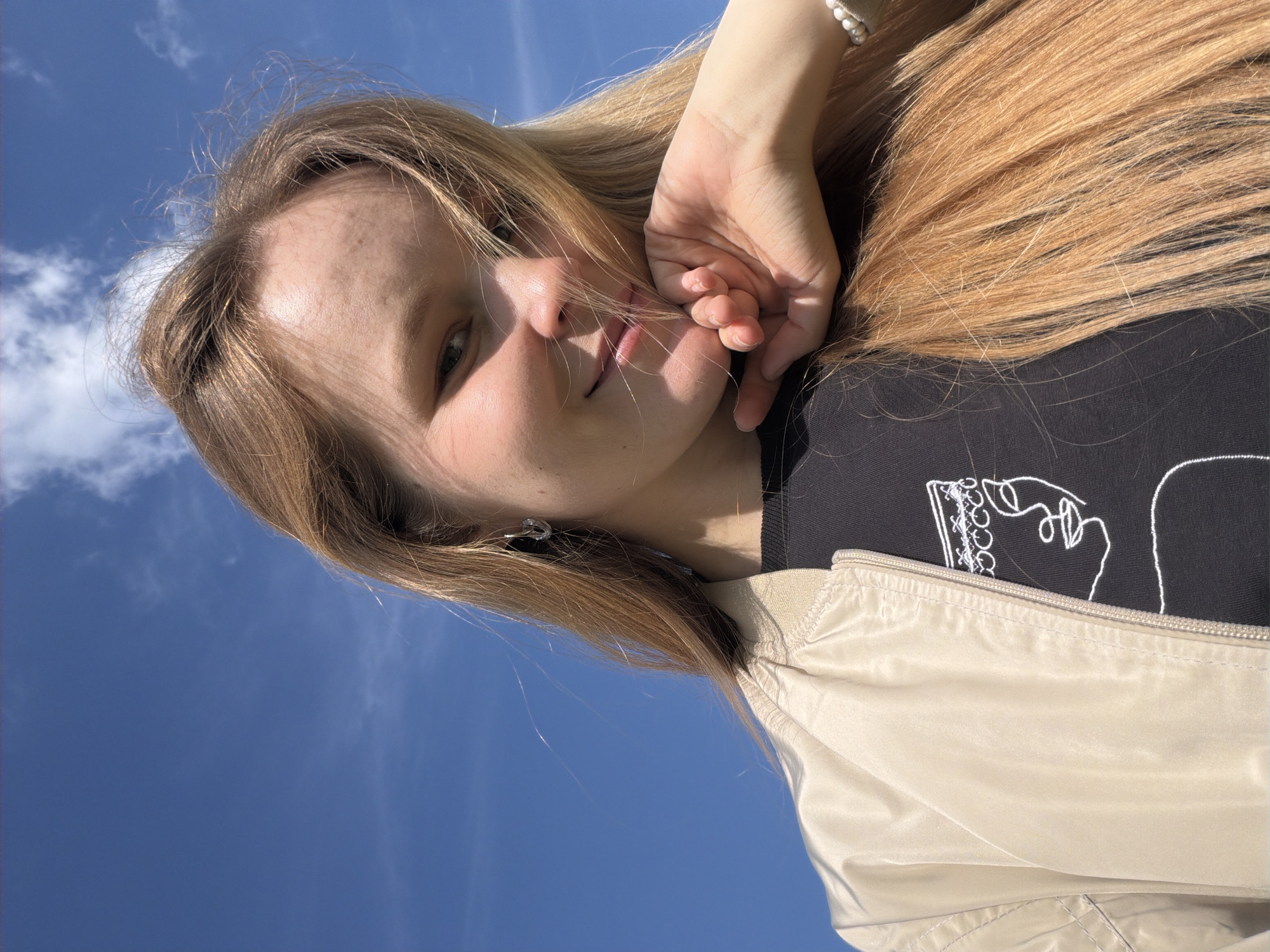 A young woman is positioned at an angle in front of a clear blue sky. She has long, blonde hair and fair skin. Her left hand is raised towards her face, with her fingers near her lips. She is wearing a black shirt with a white line drawing on it, and a beige jacket is partially visible. An earring is visible in her left ear. The location is Ильнеть, Russia.