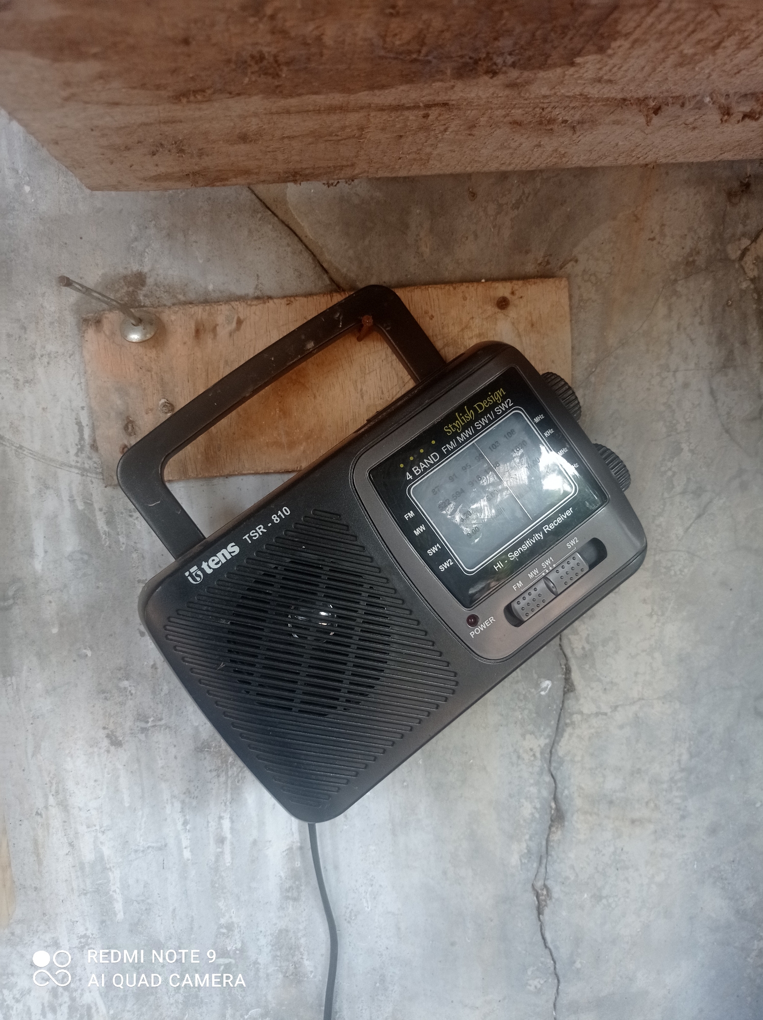 A black portable radio with a handle rests on a piece of wood which is on a concrete surface. The radio is labeled "tens TSR - 810" and "Stylish Design 4 BAND FM/MW/SW1/SW2 Hi - Sensitivity Receiver". There are knobs and a dial on the radio. The background appears to be an outdoor setting with a wooden plank above the radio. The concrete surface has cracks and is weathered. The scene is located in Cipawon, Indonesia.