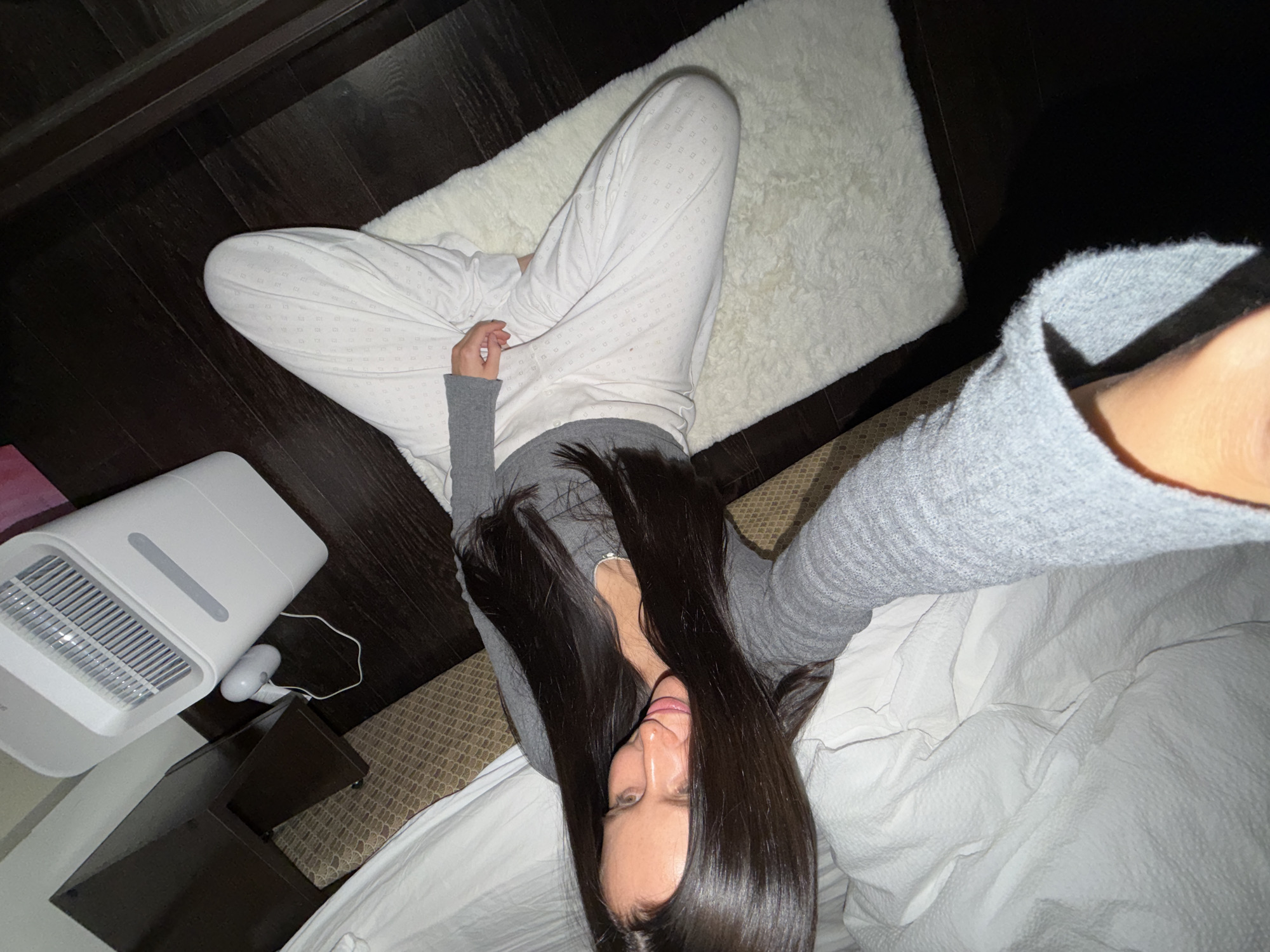 A person is lying on a bed with white linens. The person is wearing a grey long-sleeved shirt and white pants with a small square pattern. The person has long, dark hair. A white air purifier sits on a dark brown table beside the bed. A white, fuzzy rug is on the dark hardwood floor next to the bed. The location is not discernible from the image.