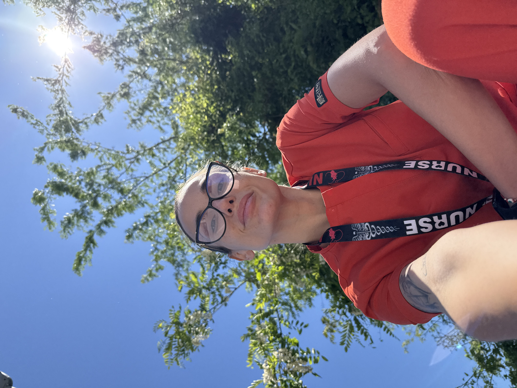 A woman with light skin and dark-framed glasses is wearing an orange scrub top with a lanyard that reads "NURSE". She is outdoors, presumably in Pleven, Bulgaria, given the prompt's request. Above her, there are tree branches with green leaves and small white flowers against a clear blue sky. The sun is shining brightly in the upper left corner of the frame. Her right arm is visible, showing a tattoo.
