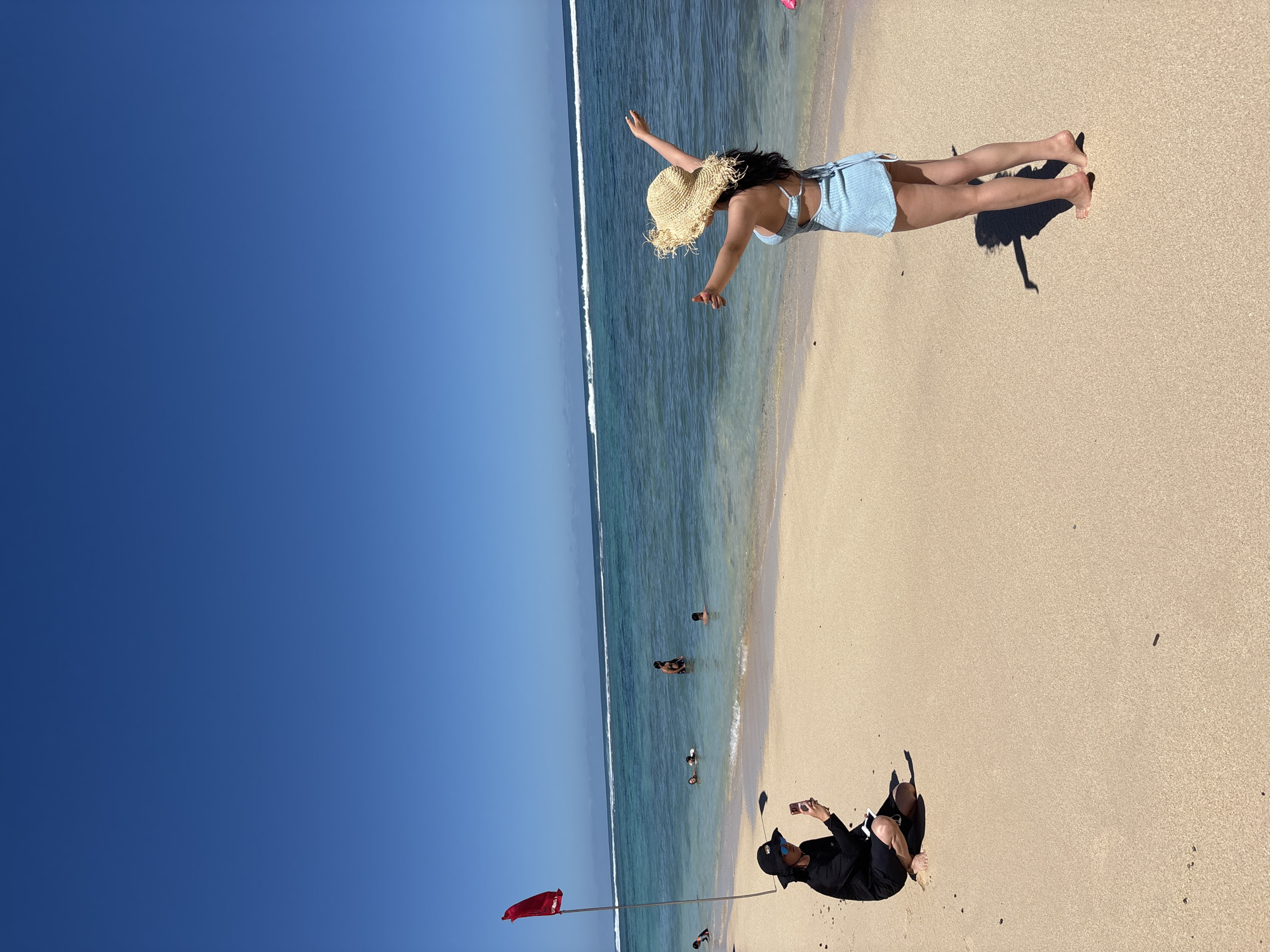 The image shows a beach in Kuta Selatan, Indonesia, under a clear blue sky. The sea is visible with small waves breaking near the shoreline. Several people are swimming in the sea. On the beach, a woman in a light blue dress and straw hat is lying down with her arms outstretched. Nearby, another individual dressed in black is sitting on the sand while looking at a phone. A red flag is planted in the sand near the water's edge. The beach itself is sandy.