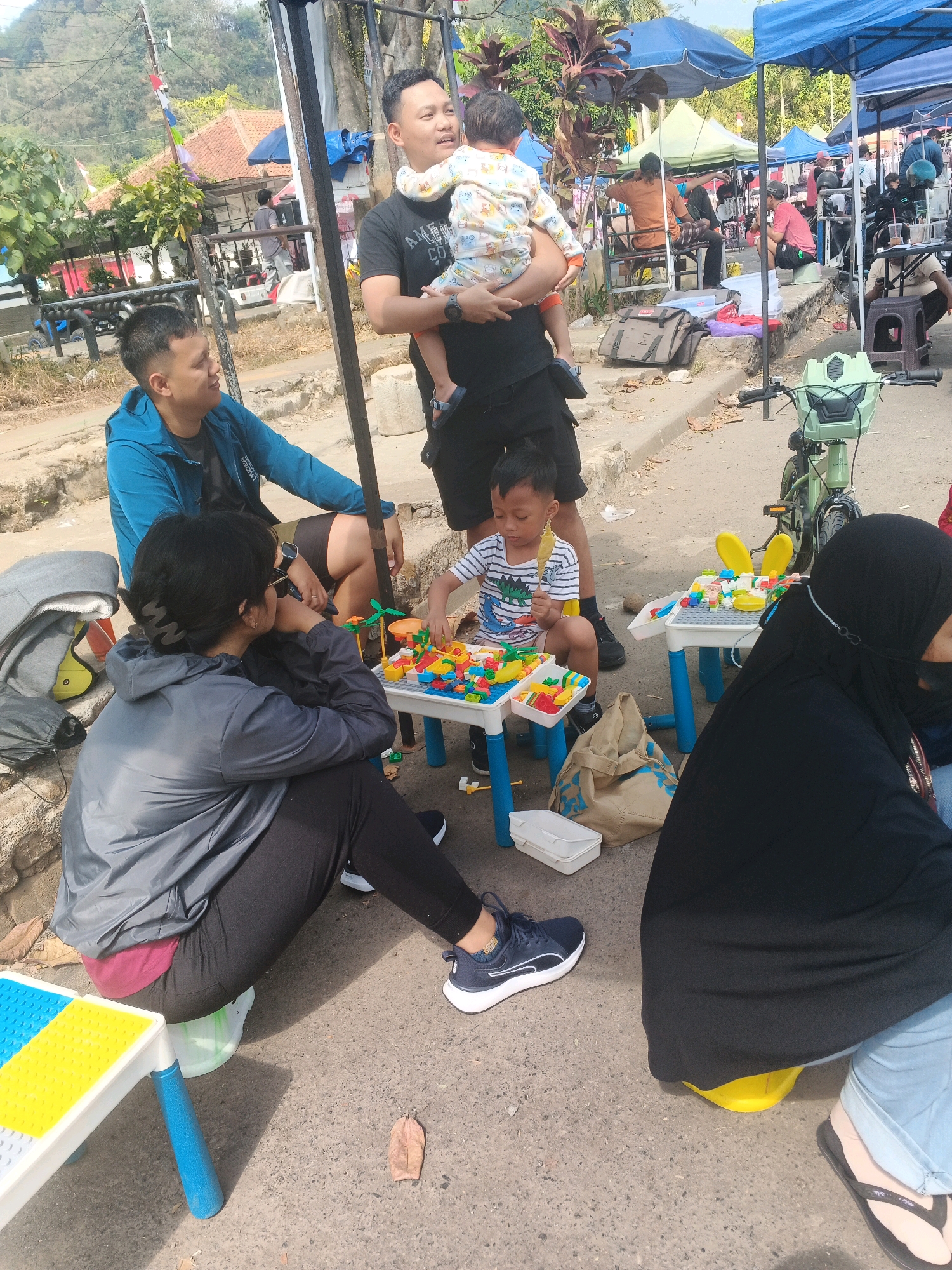 The image depicts an outdoor scene, likely in a public space within Ungaran, Indonesia, characterized by a casual gathering. In the foreground, a man holds a baby dressed in a patterned outfit. To his left, another man in a blue jacket is seated. A woman in a grey jacket and black leggings is also seated, looking to her left. A young boy in a striped shirt is sitting at a low table playing with building blocks. Another individual, wearing a black hijab and partially visible, is seated nearby. Several other people are visible in the background, some under tents, suggesting a market or event. Objects like bags, a bicycle, and a child's play table with colorful blocks are scattered around. The environment appears to be a street or pathway with trees and buildings in the distance, indicating an urban or semi-urban setting. The lighting suggests it is daytime.