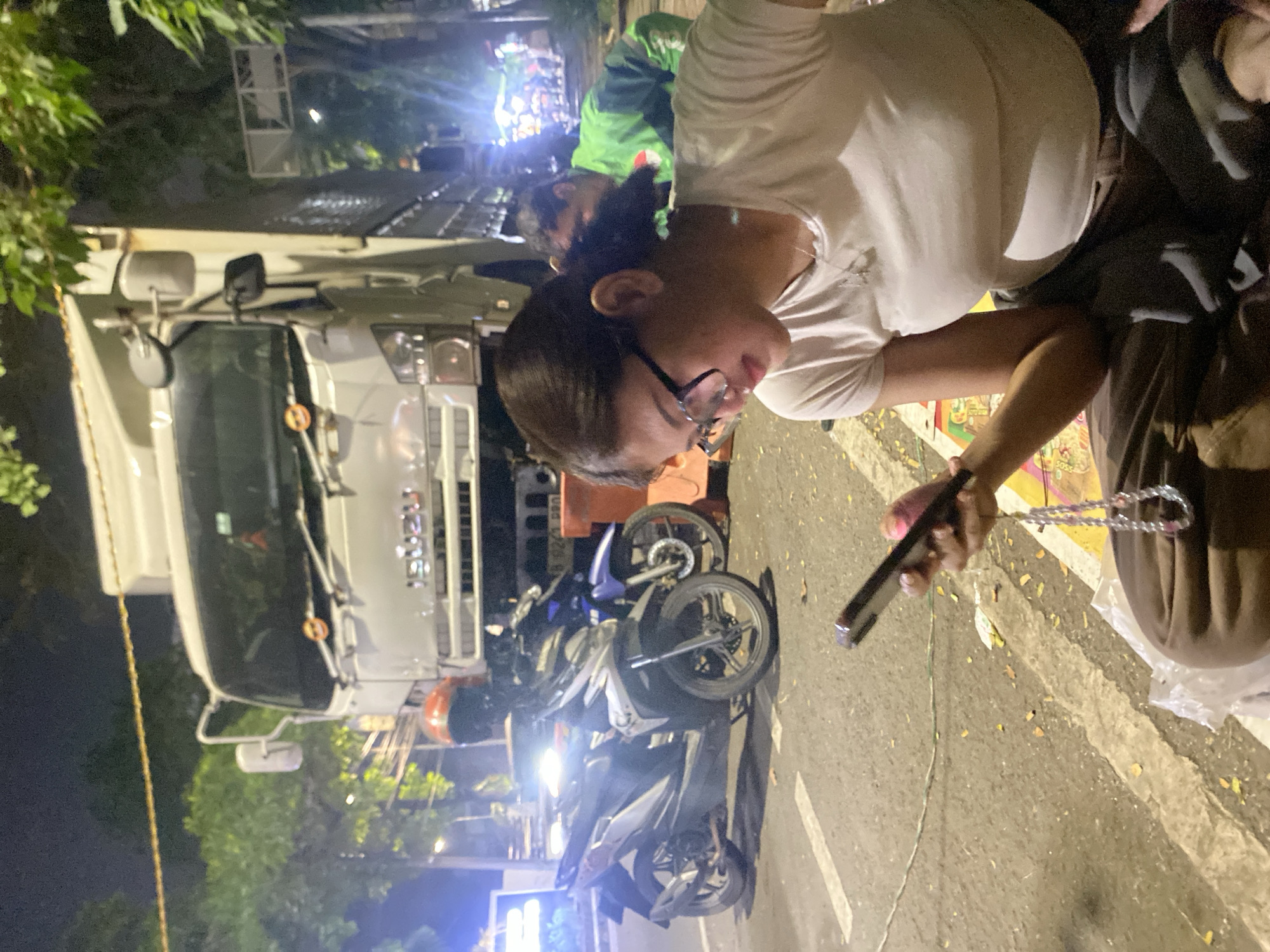 The media file shows a nighttime scene on a street in Tangerang, Indonesia. A white truck is parked on the left side of the image. Several motorcycles are parked in front of the truck, with one partially obscured by the truck's side mirror. A woman in a white t-shirt and glasses is holding a smartphone, looking down at it. She appears to be wearing a necklace. To her right, another person in dark clothing is visible. In the background, a person in a bright green jacket is standing, and there are lights and foliage. The overall lighting suggests it is dark outside, and the presence of leaves on the ground could indicate a time of year with falling leaves or recent rain. The scene appears to be a roadside, possibly an accident scene or a gathering of people with their vehicles. There is no visible text in the image.