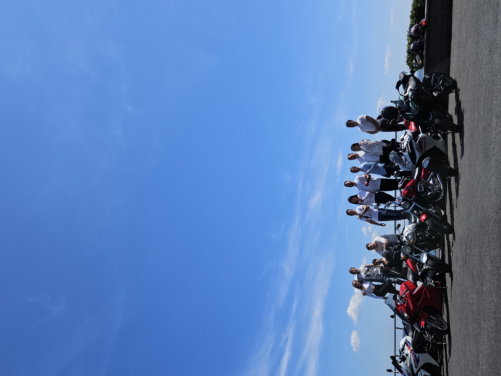 In Ulyanovsk, Russia, a group of ten individuals, comprising six men and four women, poses for a photograph alongside a line of motorcycles. The group is positioned on a raised area, possibly a viewing platform or embankment, overlooking a body of water, suggested by the distant blue expanse under a clear sky with wispy clouds. The motorcycles are parked in front of them, creating a colorful array of red, white, and black vehicles. The people are casually dressed, some standing and others leaning on or near the motorcycles. The scene suggests a gathering or outing, possibly a motorcycle club meeting or a recreational ride.